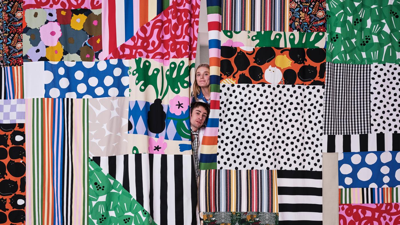 Two people stick their heads out between two patchwork curtains made from fabrics from the Tyg collection.