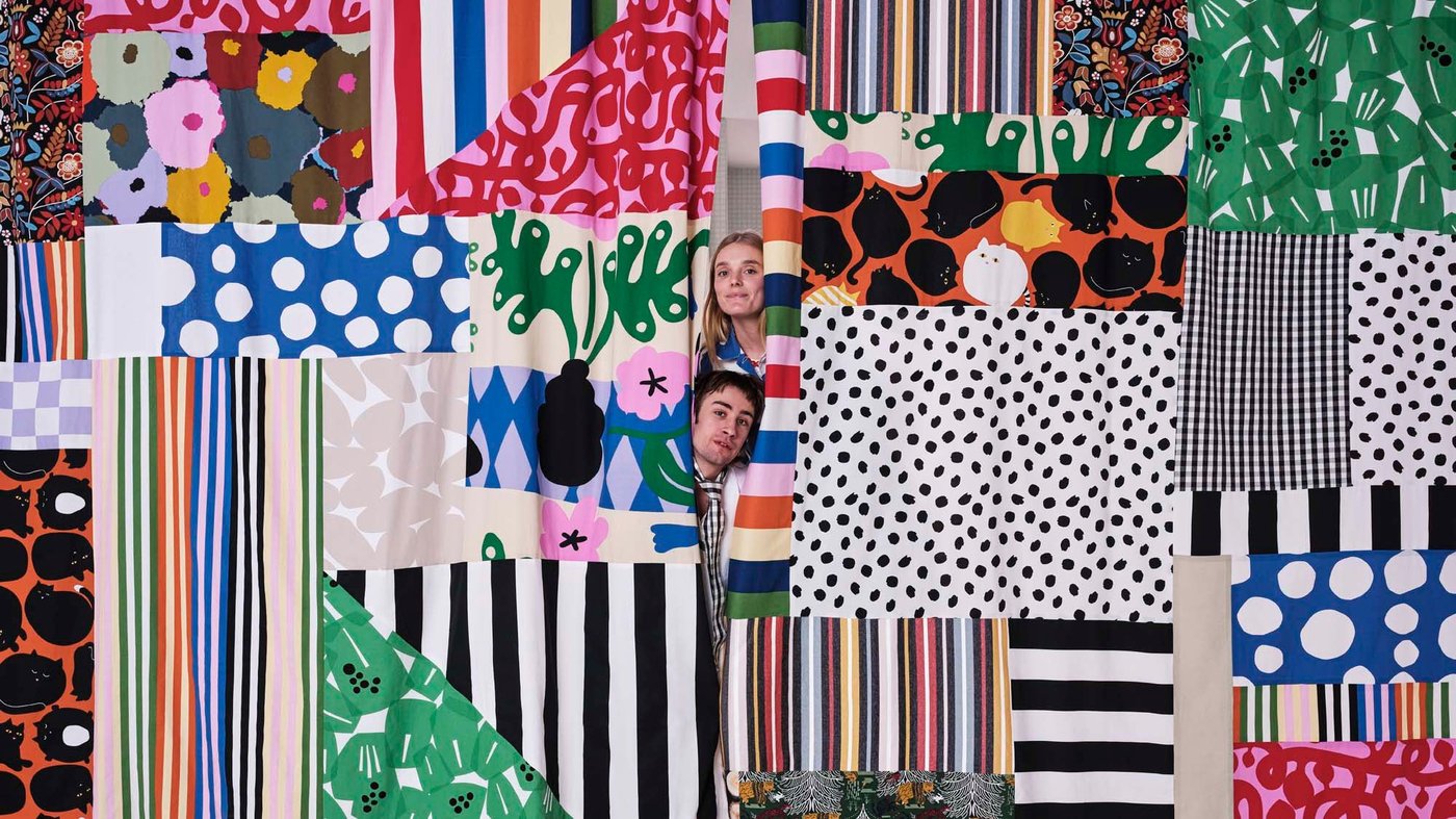 Two people stick their heads out between two patchwork curtains made from fabrics from the Tyg collection.