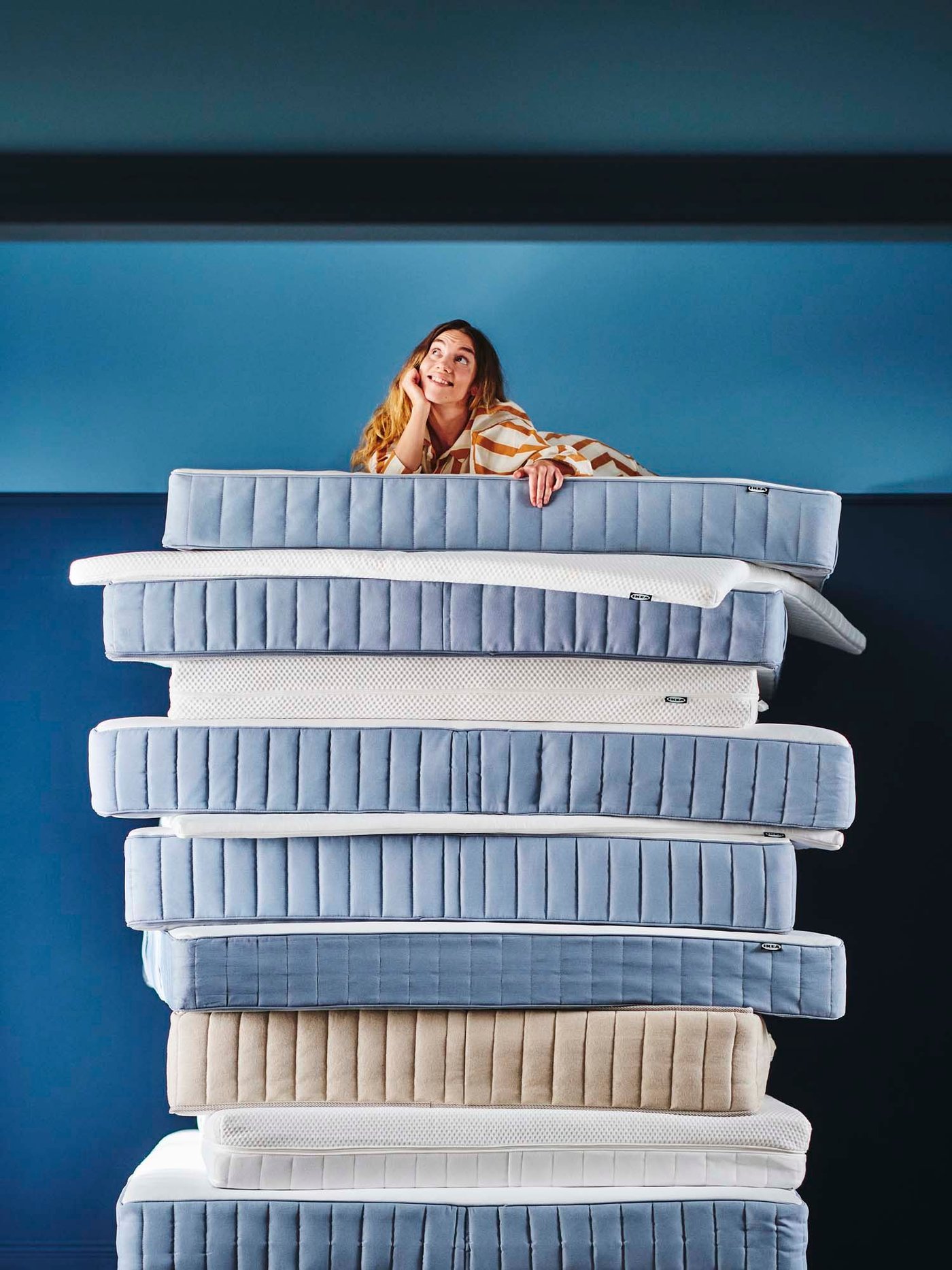 A person leans on a large stack of mattresses against a blue background.