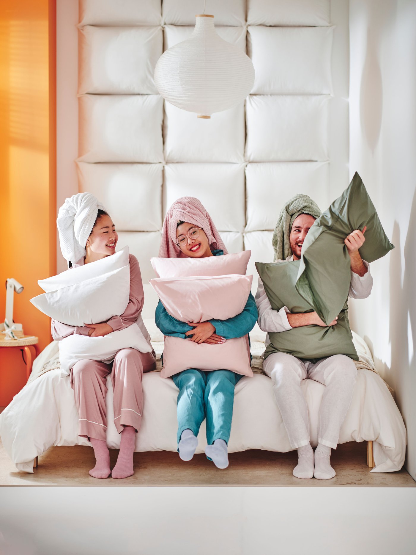 In a room with pillow on its walls, three smiling people are sitting next to each other on a bed, with one pillow each in their hands, dressed with towels on their heads.