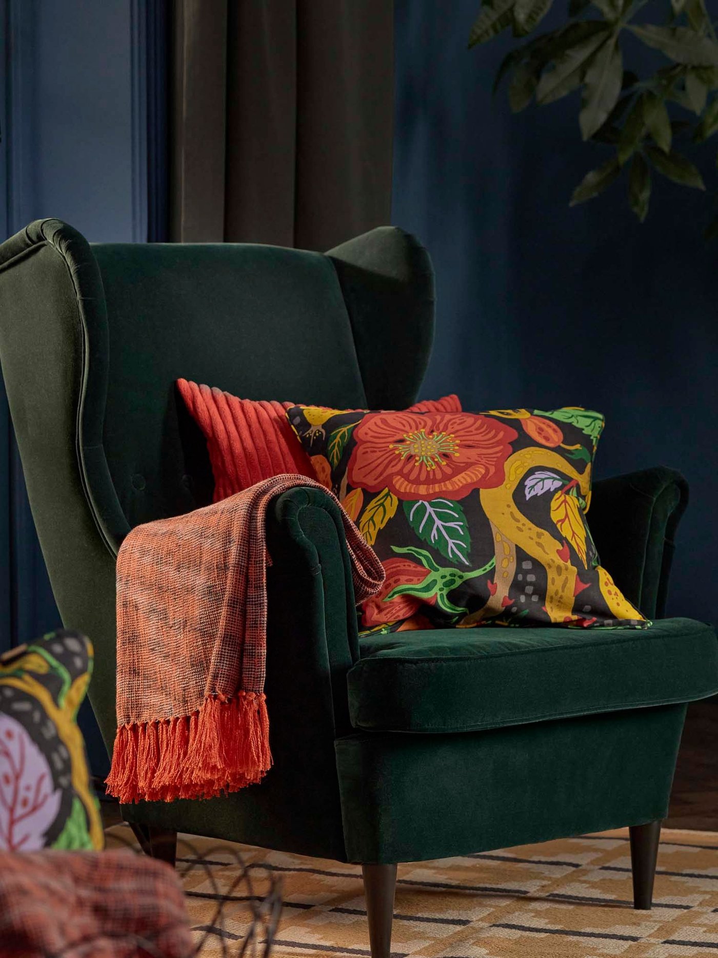 A room with an orange HÖSTAGILLE throw and two cushions in HÖSTAGILLE cushion covers on a dark green STRANDMON wing chair.