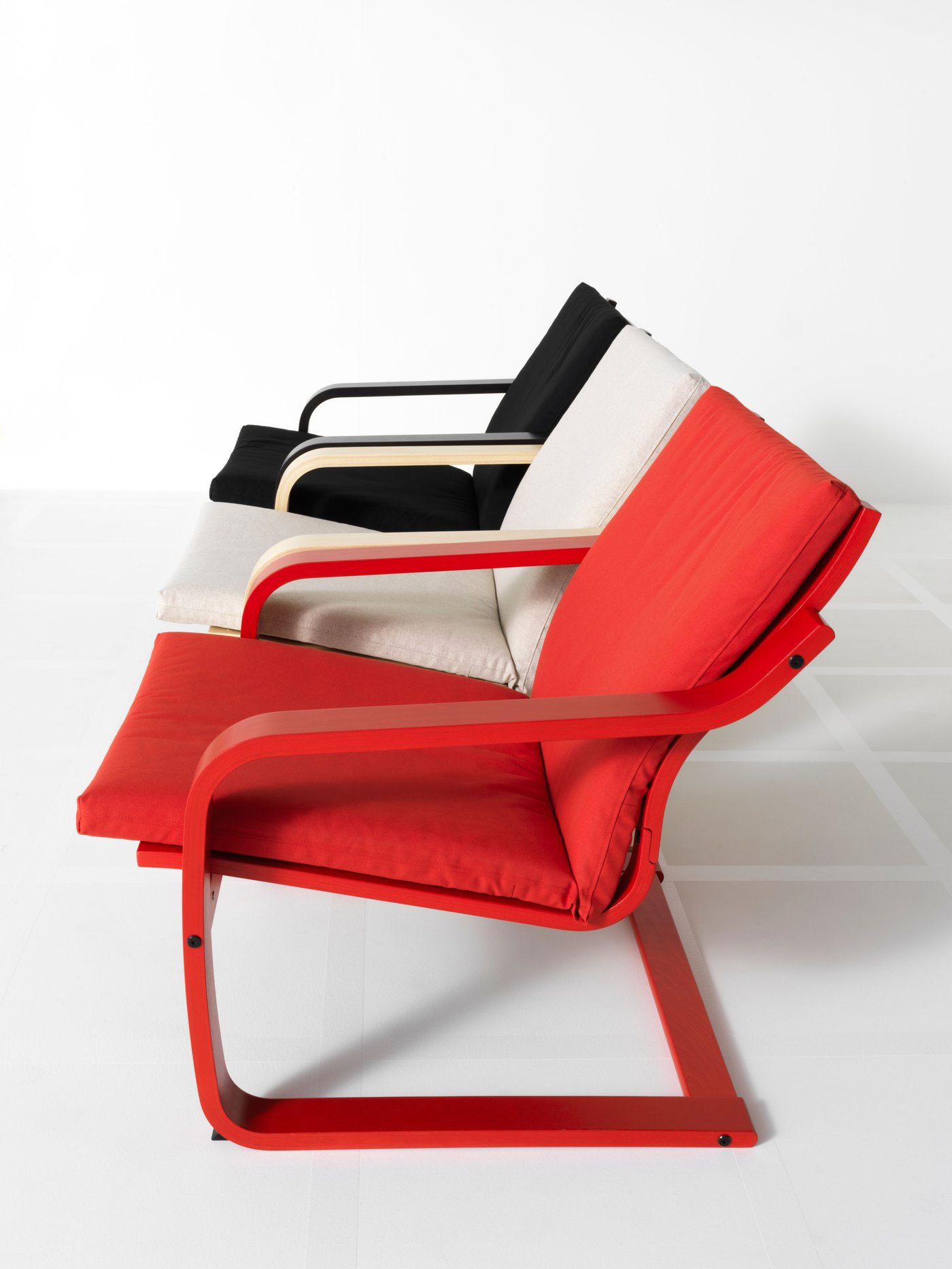 A red, white and black POÄNG chair are standing next to each other in a row, seen from the side.