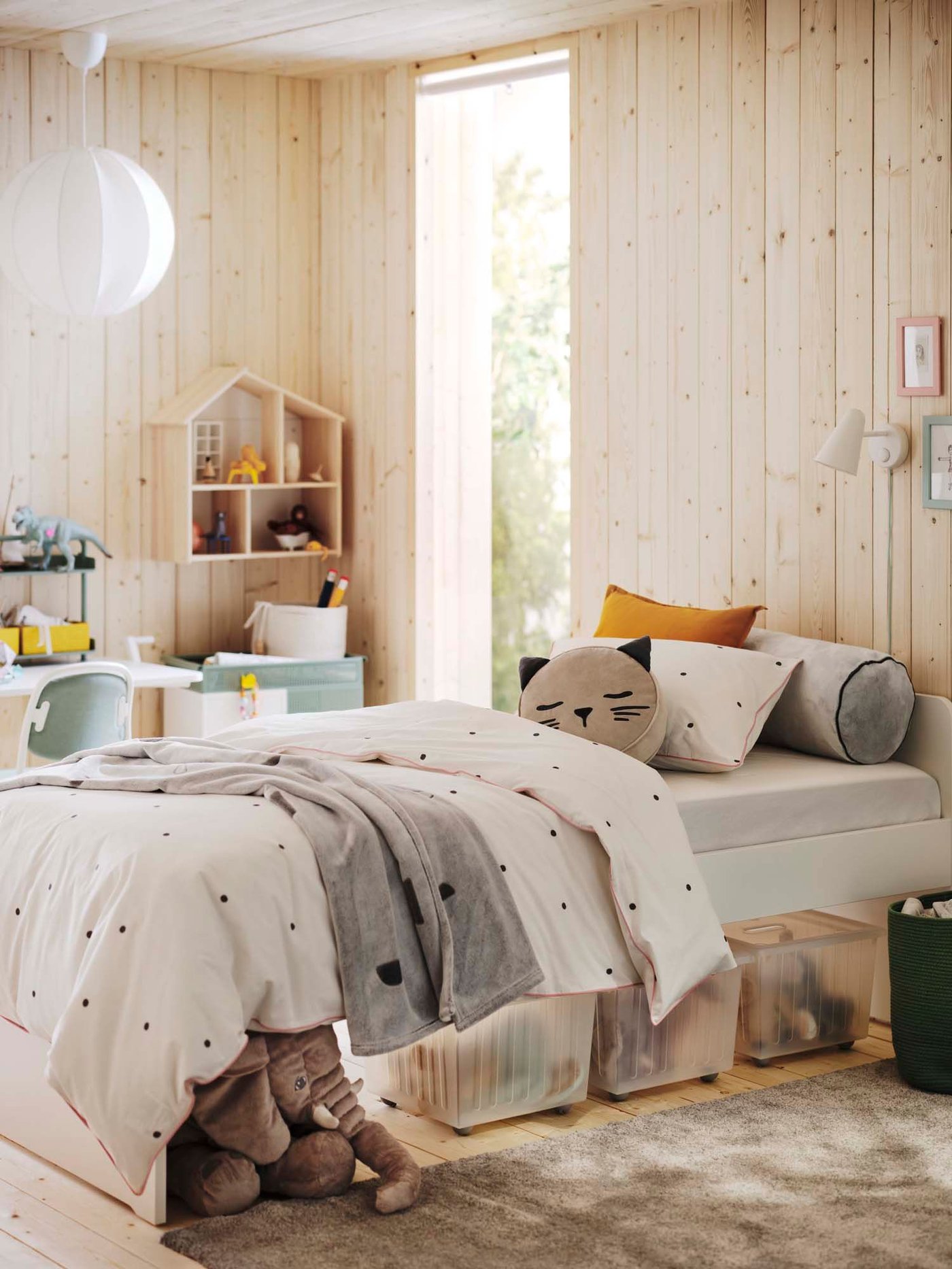 A child's bedroom with light wooden walls, a bed with dotted bedding, and a cat pillow. VESSLA storage boxes and a plush elephant are under the bed.