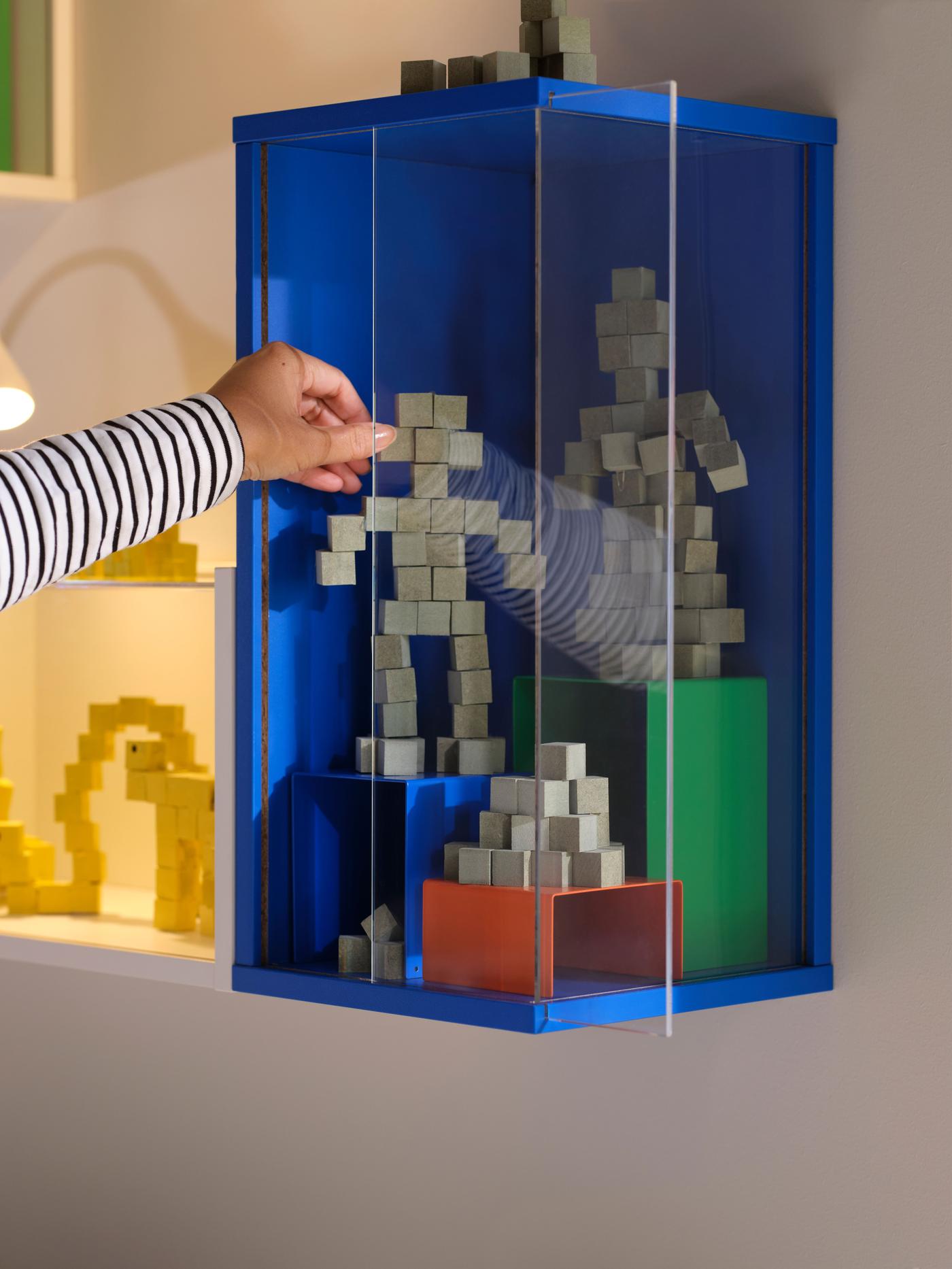 From the side, a hand is reaching forward to touch figurines and trophies standing in a blue wall-mounted cabinet
