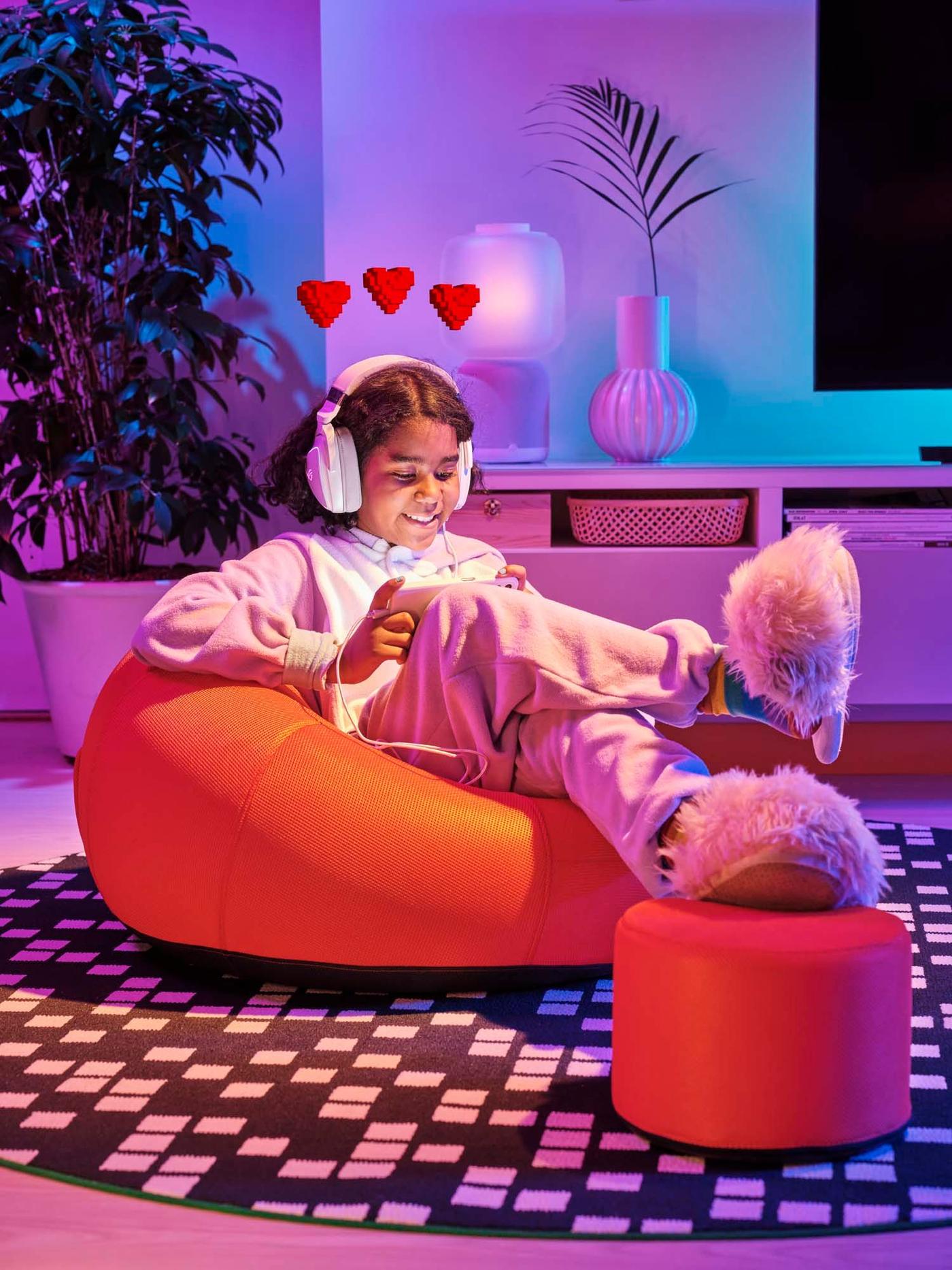 A girl sits in a low seat from the BRÄNNBOLL collection. She has her feet up on a footstool. She is wearing headphones.