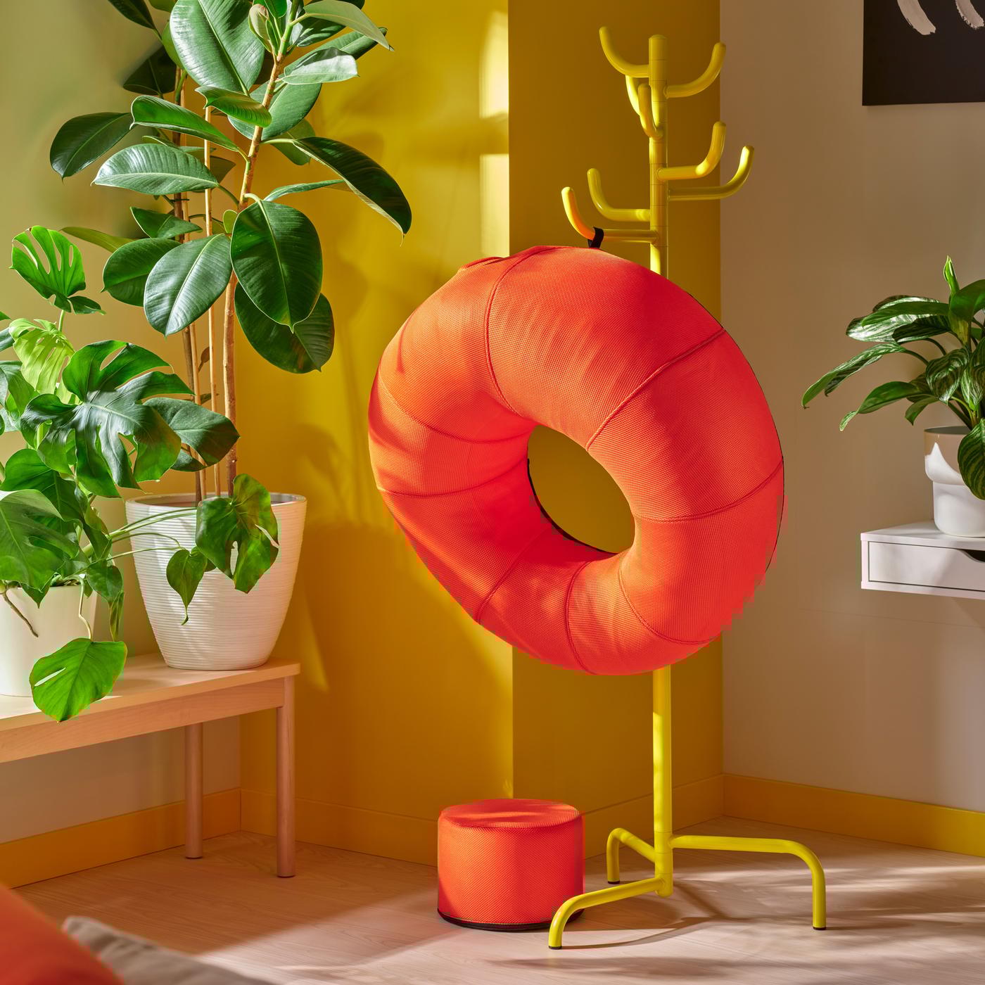 A red inflatable donut chair is hanging on a yellow hanger in a bright room with plants