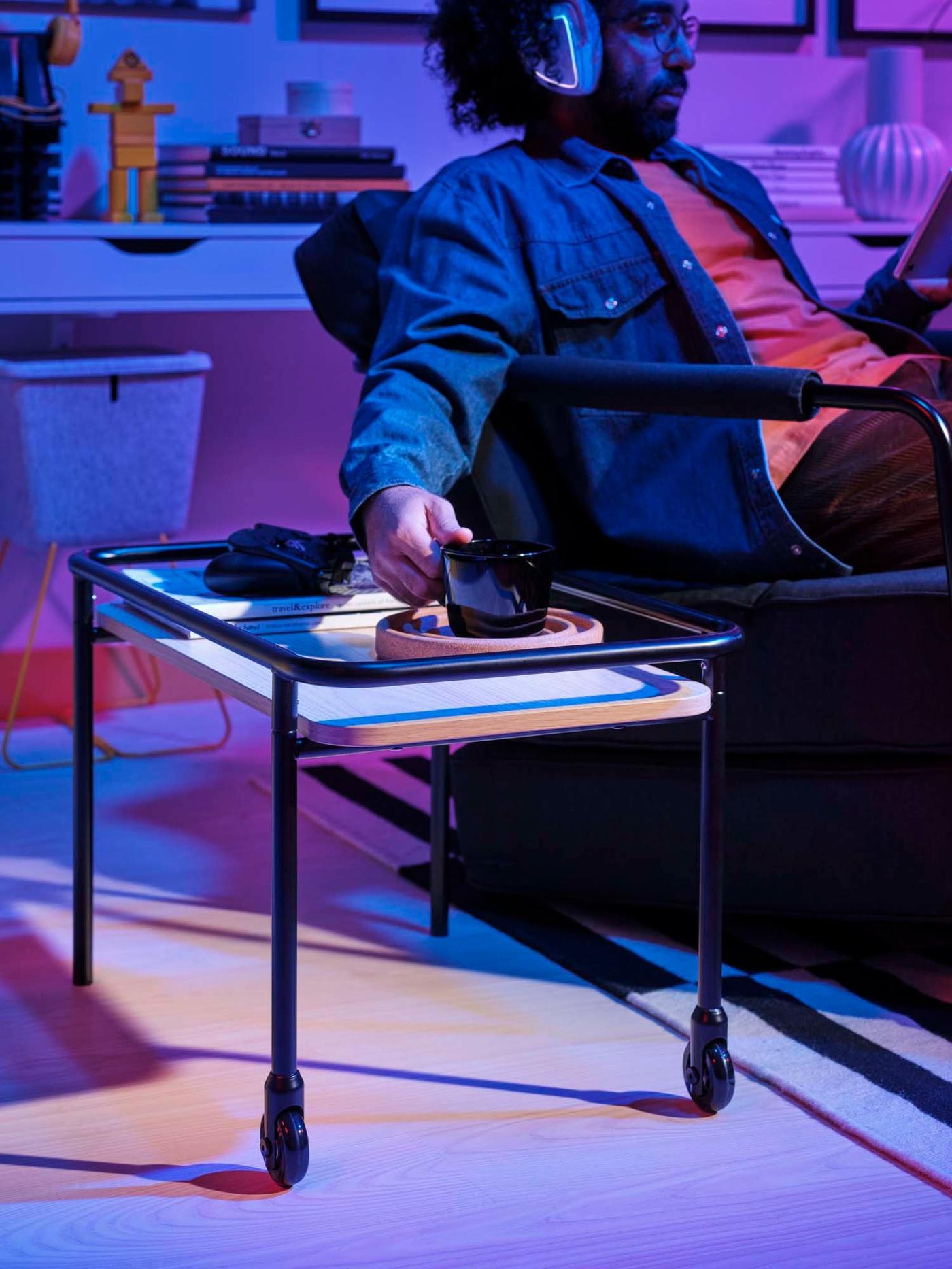 A man sits in an armchair. He is taking something from the side table on wheels.