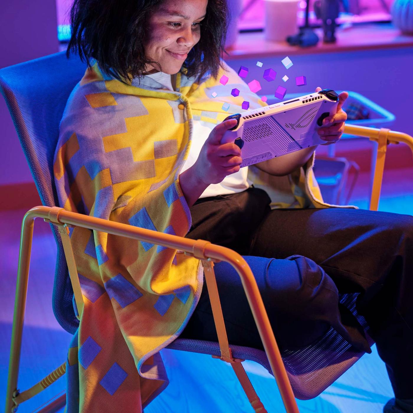 A girl is sitting in a dark room, in a yellow and beige easy chair, with a yellow blanket wrapped around her neck. She is smiling towards the bright coloured screen that she is holding in front of her.