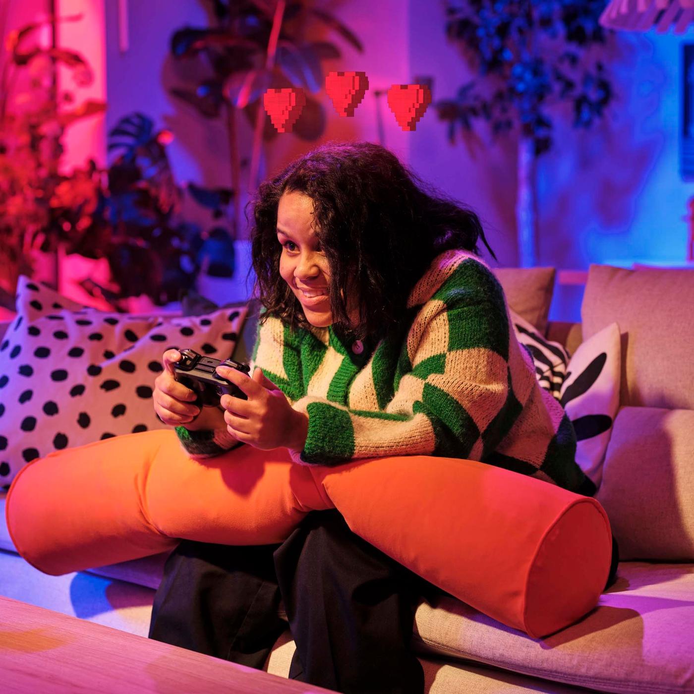 A girl sits on a sofa playing a video game. On her lap is a long orange bolster cushion from the BRÄNNBOLL gaming collection.