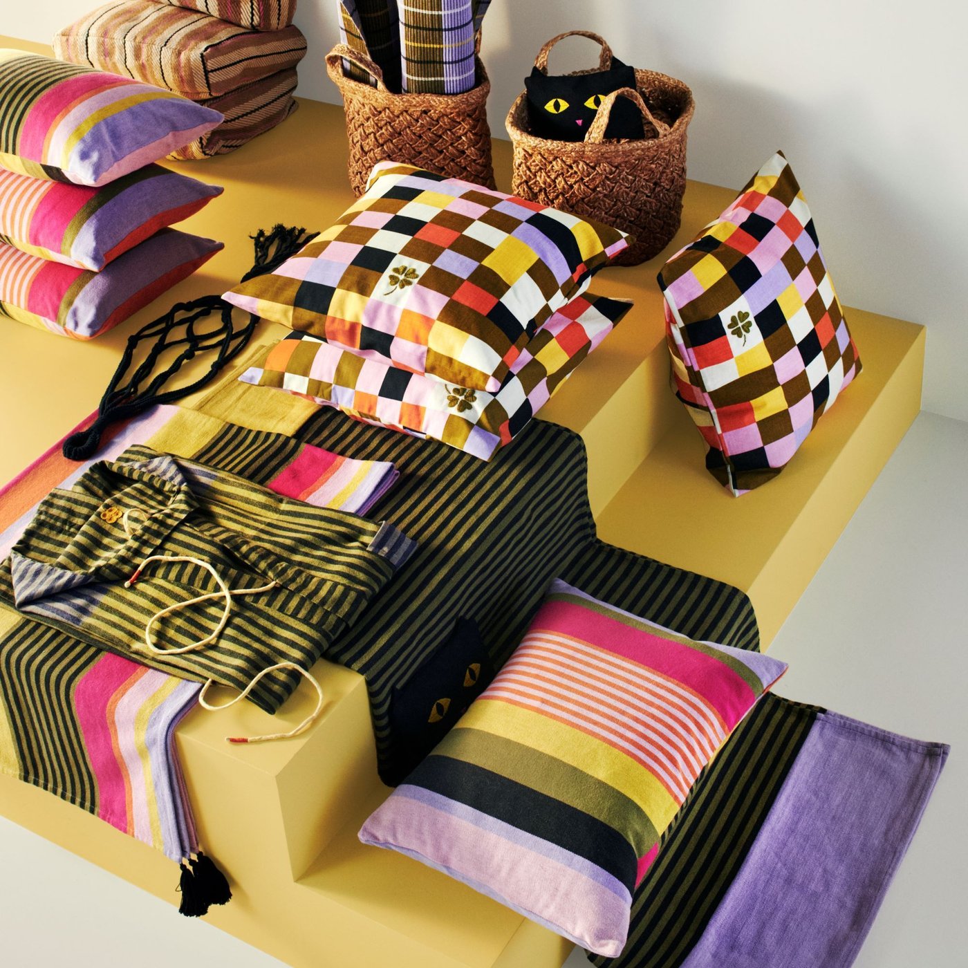 An aerial image of products from the MÄVINN collection, showing colourful cushions with square and stripe patterns and woven baskets.
