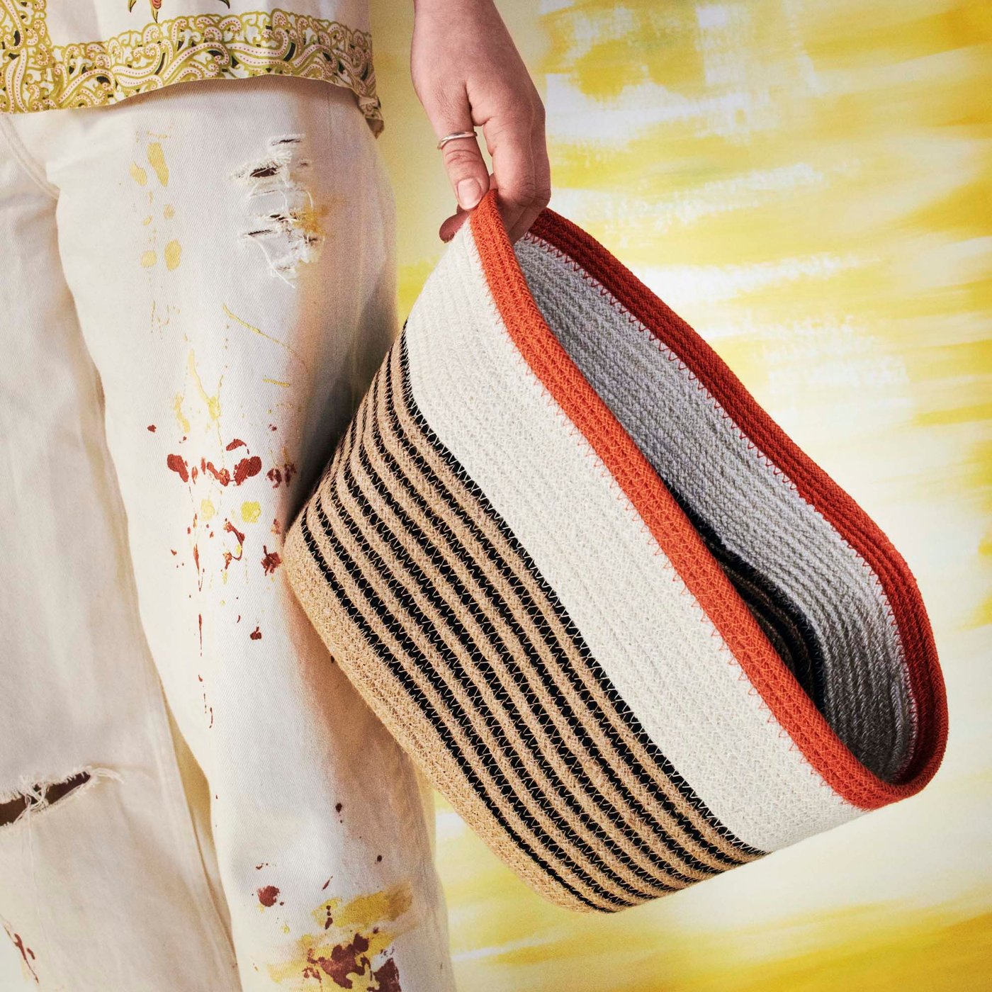 The legs and hand of a person holding a striped multicolour MÄVINN basket by its rim.