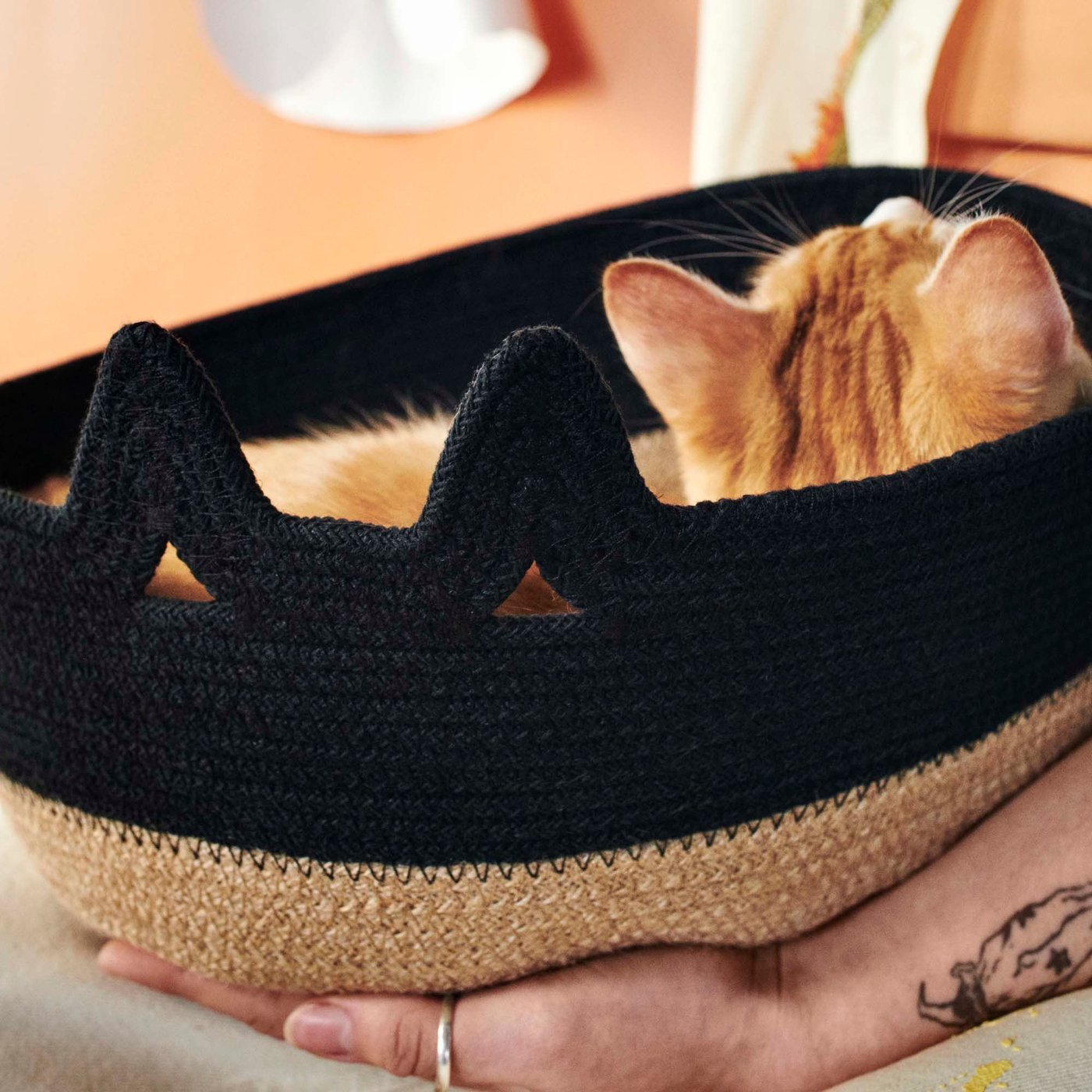 A hand holding a ginger cat in a black/natural MÄVINN basket.