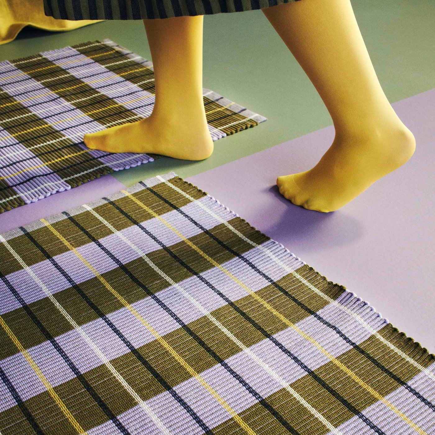 Feet in yellow tights walking across two striped multicolour MÄVINN rugs.