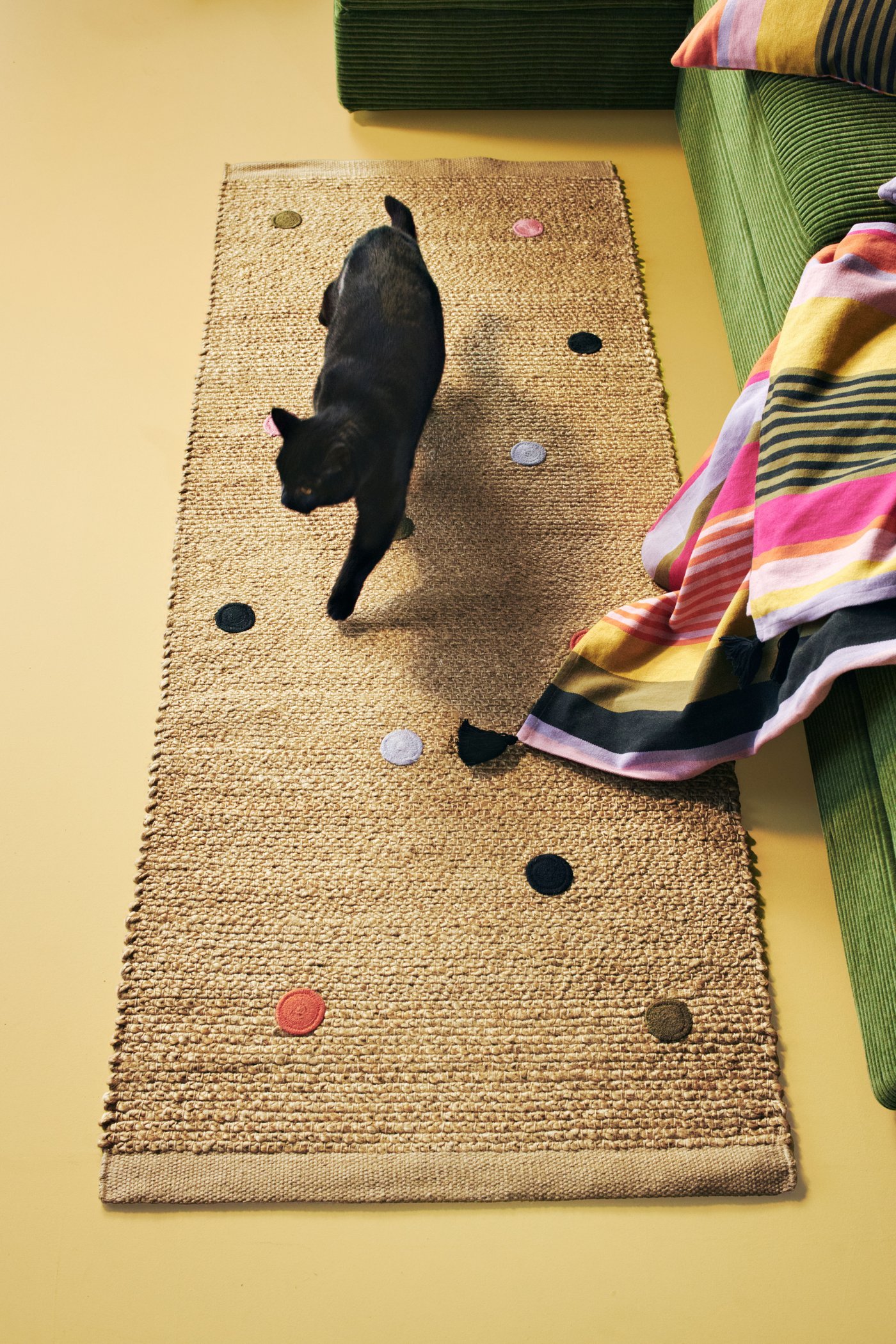 A black cat is crawling on a woven rug on the yellow colored floor. Besides there is a green colored bed and a colorful striped throw on it.