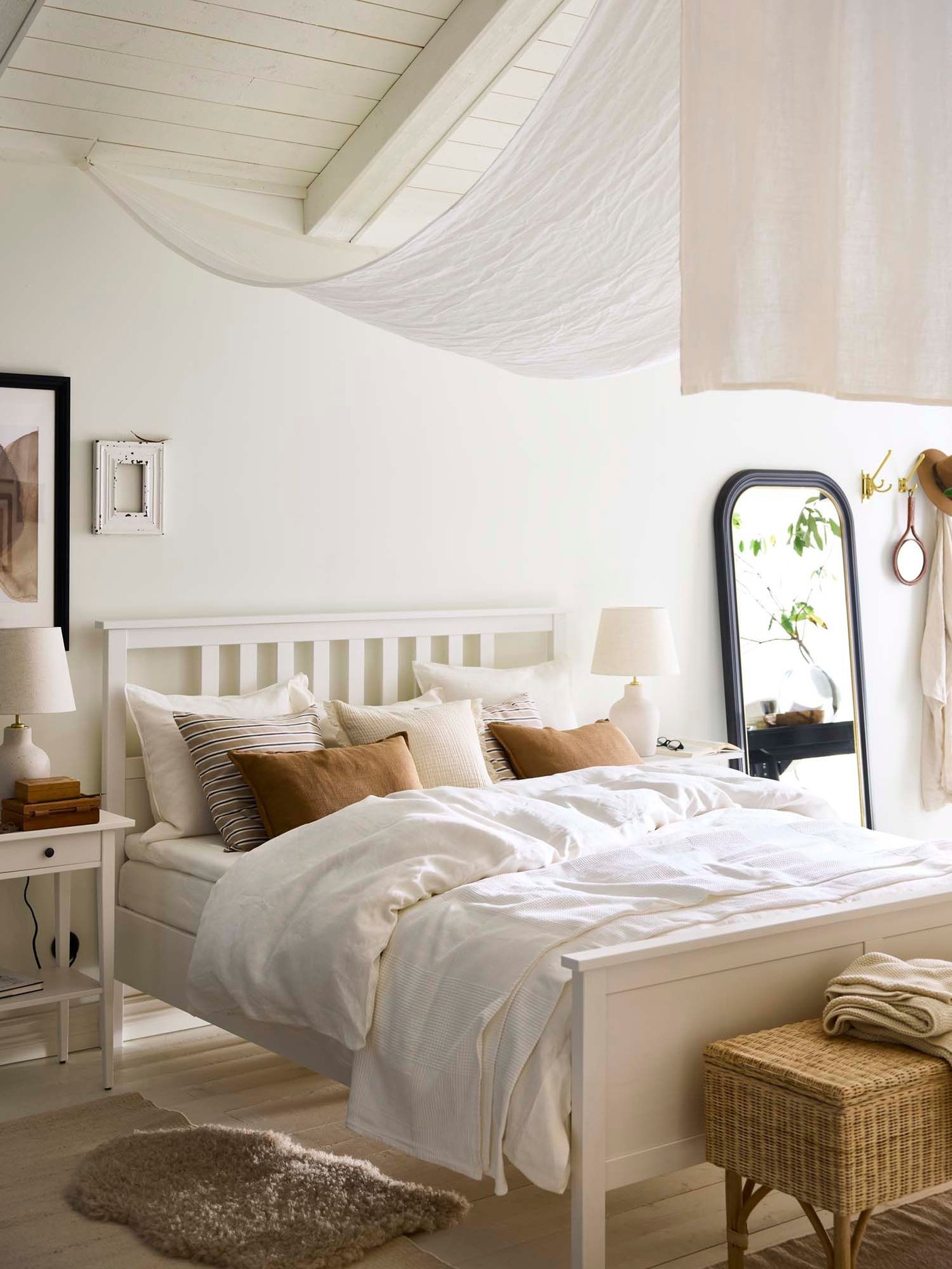 A white bedroom with coloured cushions on a white wood bed and light muslin fabric hanging from the ceiling.