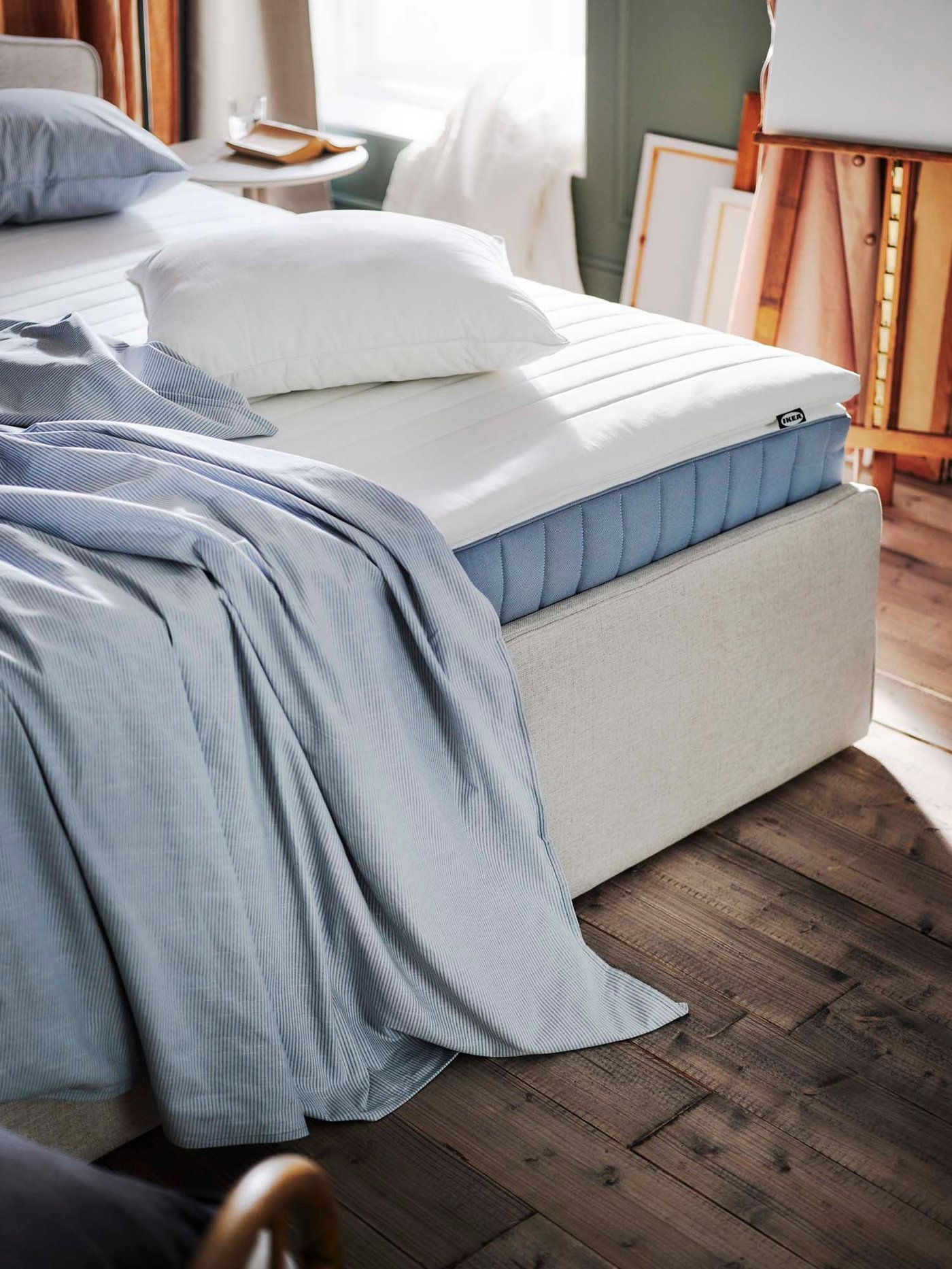 The corner of a bed, showing the mattress, with bedlinen folded and draped over the edge.