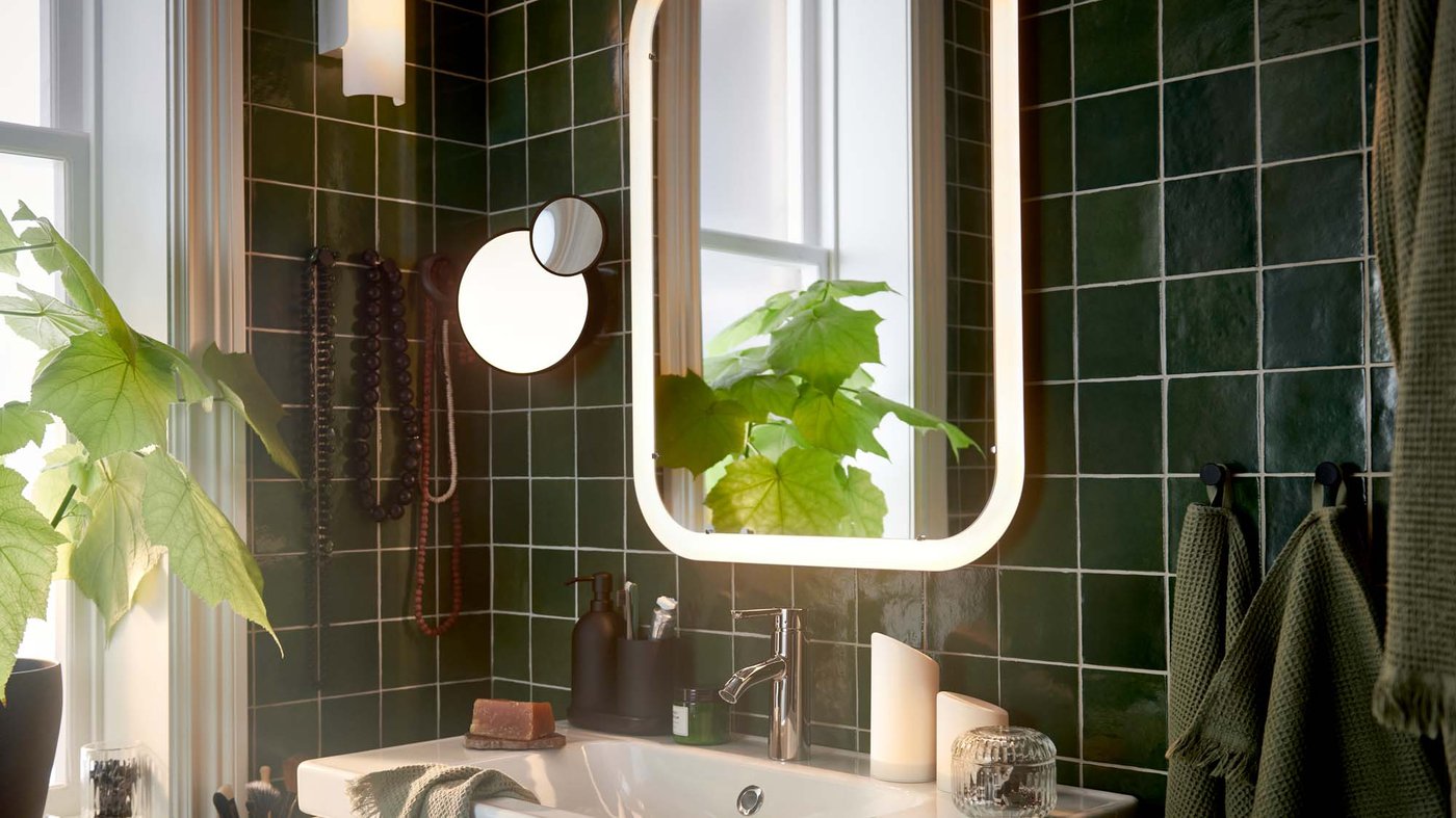 The corner of a green tiled bathroom with a leafy potted plant reflected in an illuminated STORJORM mirror.