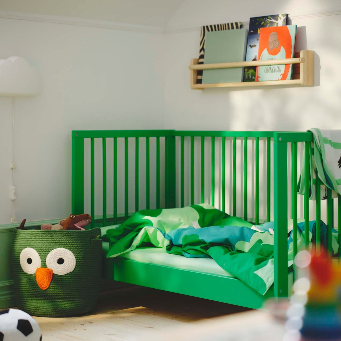 In a toddler room, a green cot sits in the corner, next to a green UPPSTÅ storage bag. Above is a bookshelf filled with children's books.