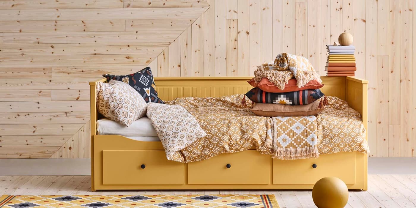 A yellow HEMNES daybed is adorned with patterned pillows, a blanket, and a stack of books. It sits on a geometric-patterned rug in front of a wood-paneled wall.