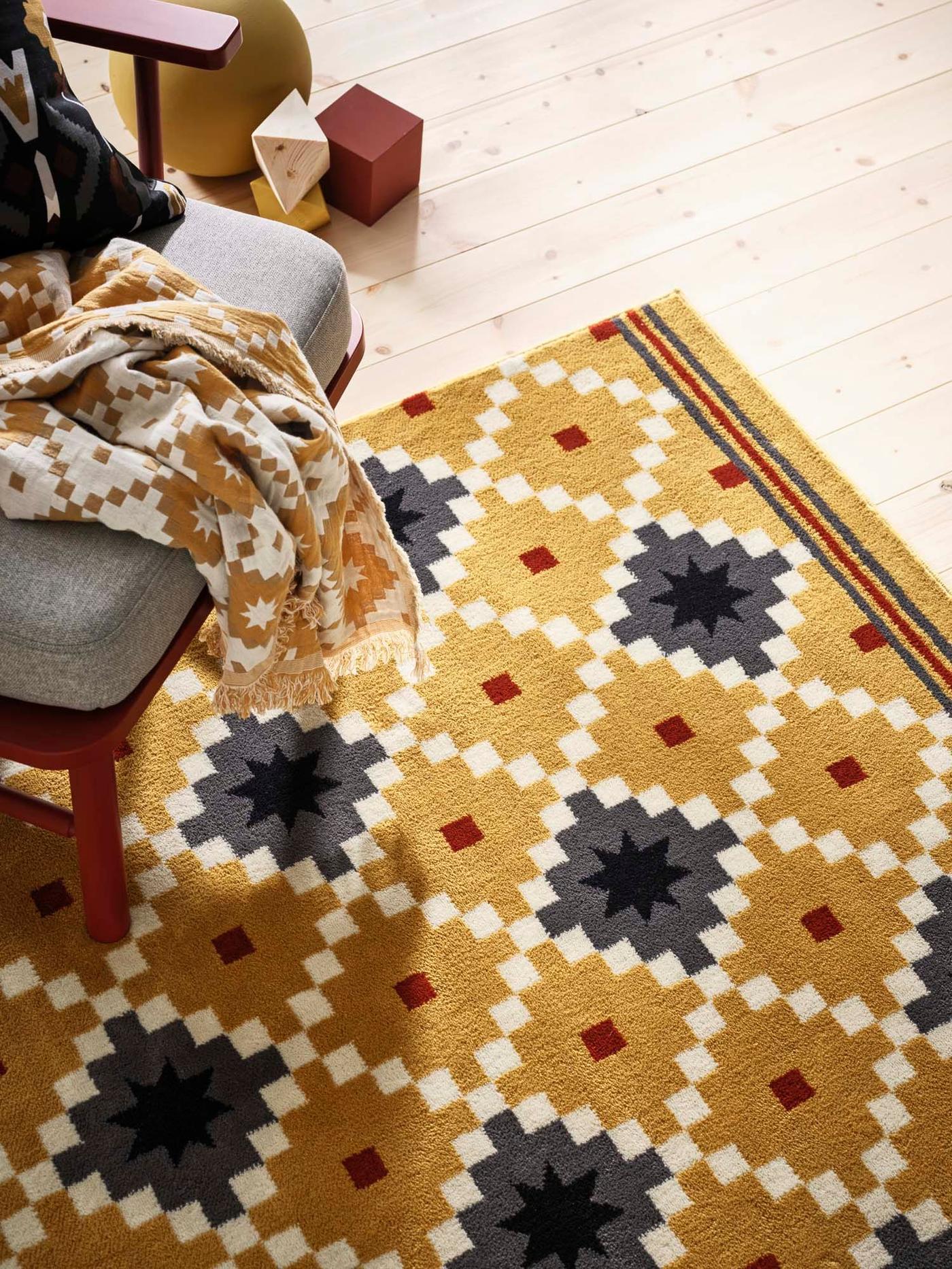 The corner of a chair sits on a patterned rug with a black and yellow geometric pattern on a wooden floor. 