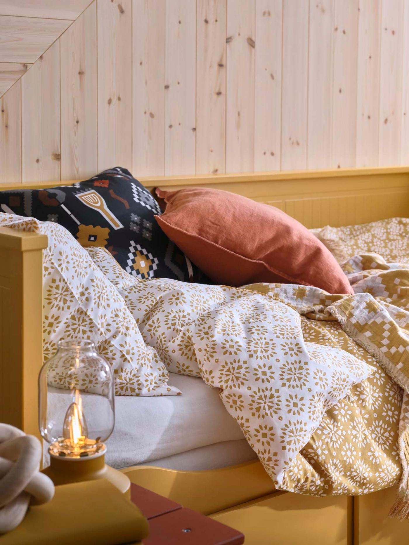 A yellow HEMNES day bed made up in patterned bedlinen, with cushions and a yellow lantern by the side of the bed.