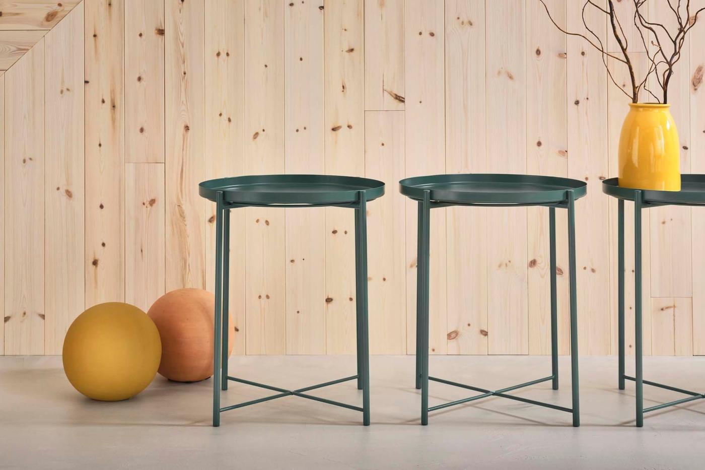 Three green side tables in a row against a wood wall backdrop. One has a yellow vase on top.