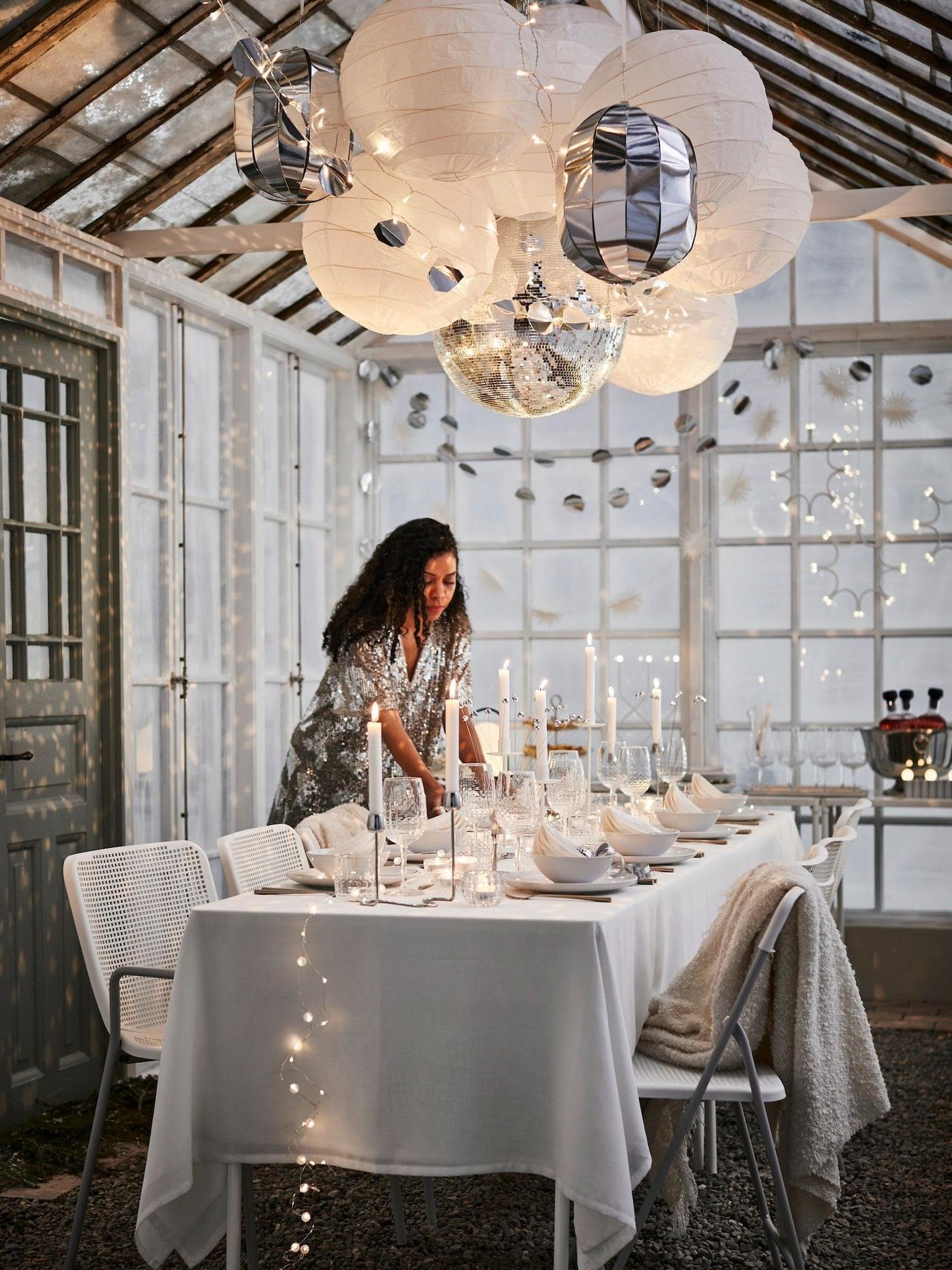 A person sets a long table inside a glasshouse decorated for a celebration in silver and white, with a cluster of REGOLIT pendant lamp shades and FRÖJDA hanging decorations suspended above the table.