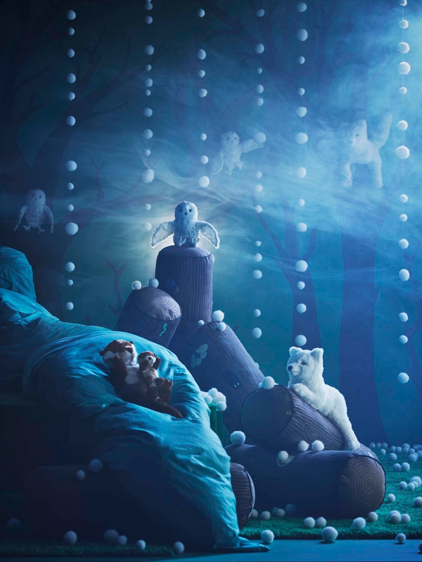 A dark and snowy scene created with the SKOGSDUVA collection. White soft balls hanging from the ceiling as matter of snow. The scene is filled with the SKOGSDUVA soft toys and cushions like the arctic fox, owl, log and otters.