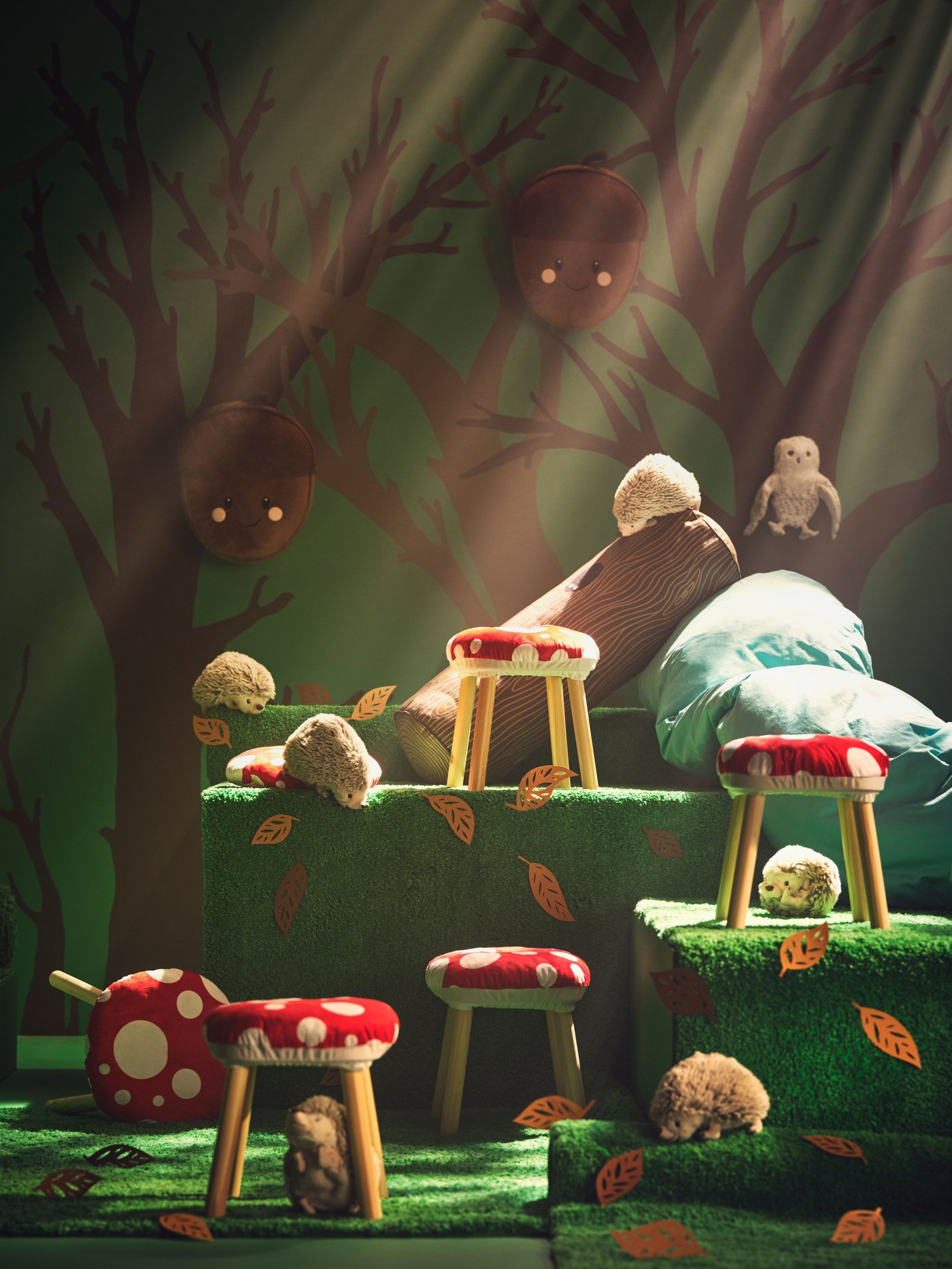 A green scene with green mats on the ground and tree paintings in the background. On the green mats red and white toadstools, hedge hocks, owls and acorn cushions hanging. 