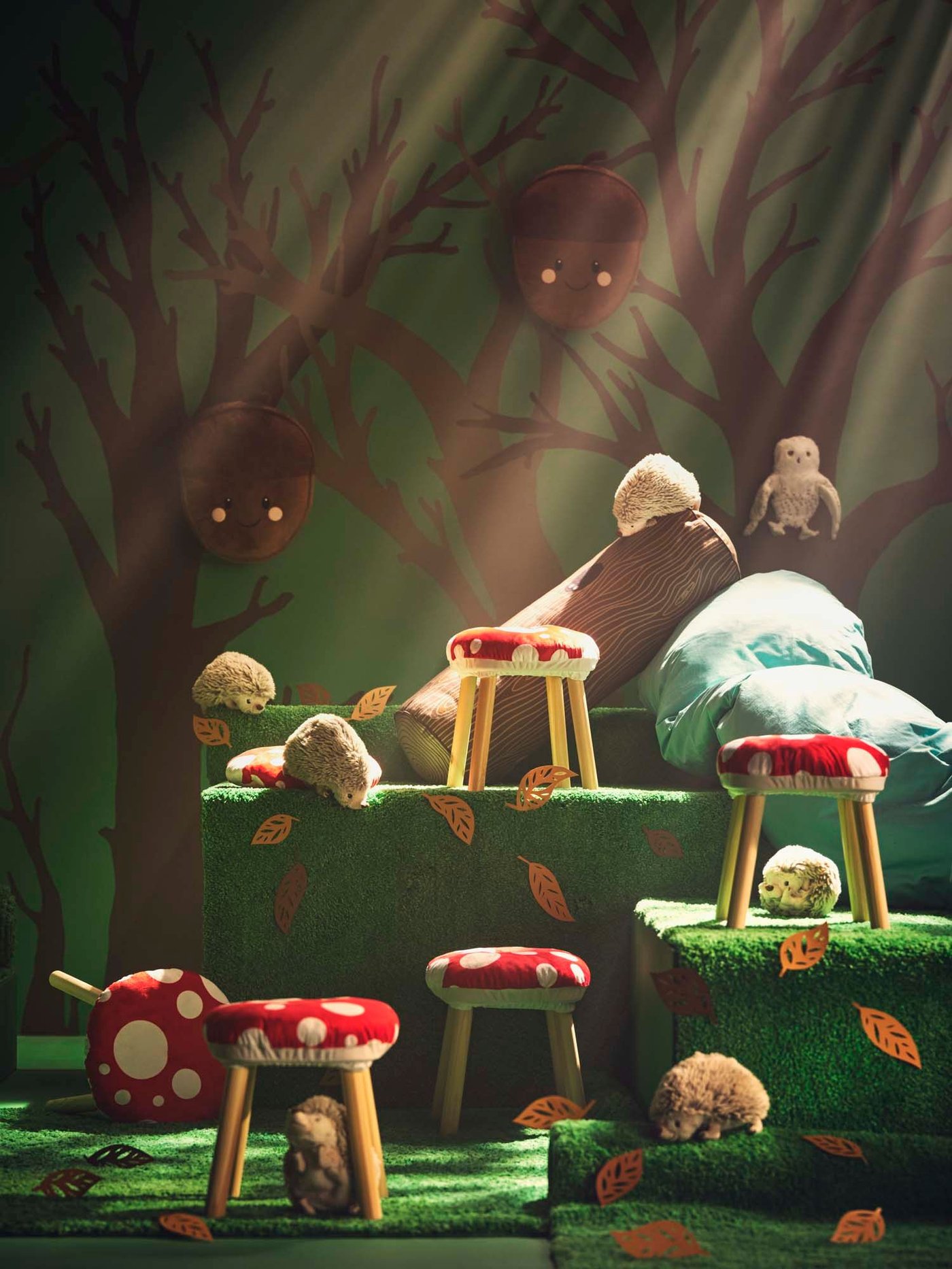 A green scene with green mats on the ground and tree paintings in the background. On the green mats red and white toadstools, hedge hocks, owls and acorn cushions hanging.