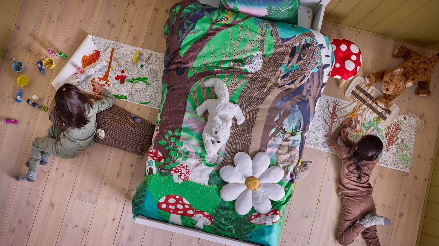 A children's bedroom with a bed. On the bed the SKOGSDUVA duvet covers, the arctic fox and the flower pillow. Underneath the bed there is a coloring page that is sticking out on both sides of the bed. On each side, children are painting the coloring page.