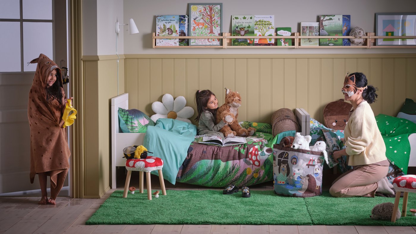 A children's bedroom with two small children and a mother surrounded by the SKOGSDUVA collection consisting of soft toys, duvets, cushions and more, all inspired by the Swedish forest and wildlife.
