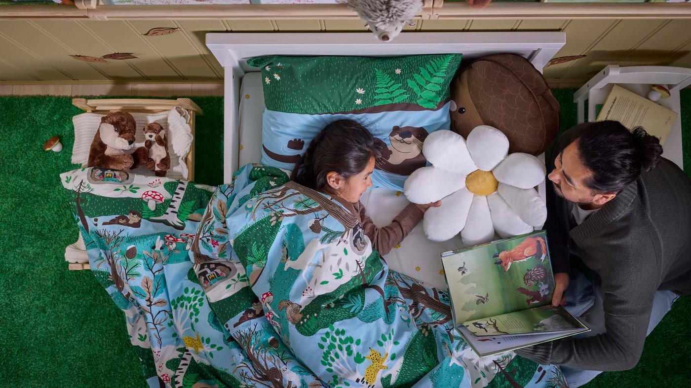 A father reading a bedtime story to his daughter by her bed. The child lays in bed under a duvet covered with the SKOGSDUVA duvet covers. In her bed the SKOGSDUVA flower pillow and the acorn pillow. Next to her bed a tiny bed with two soft toy otters.