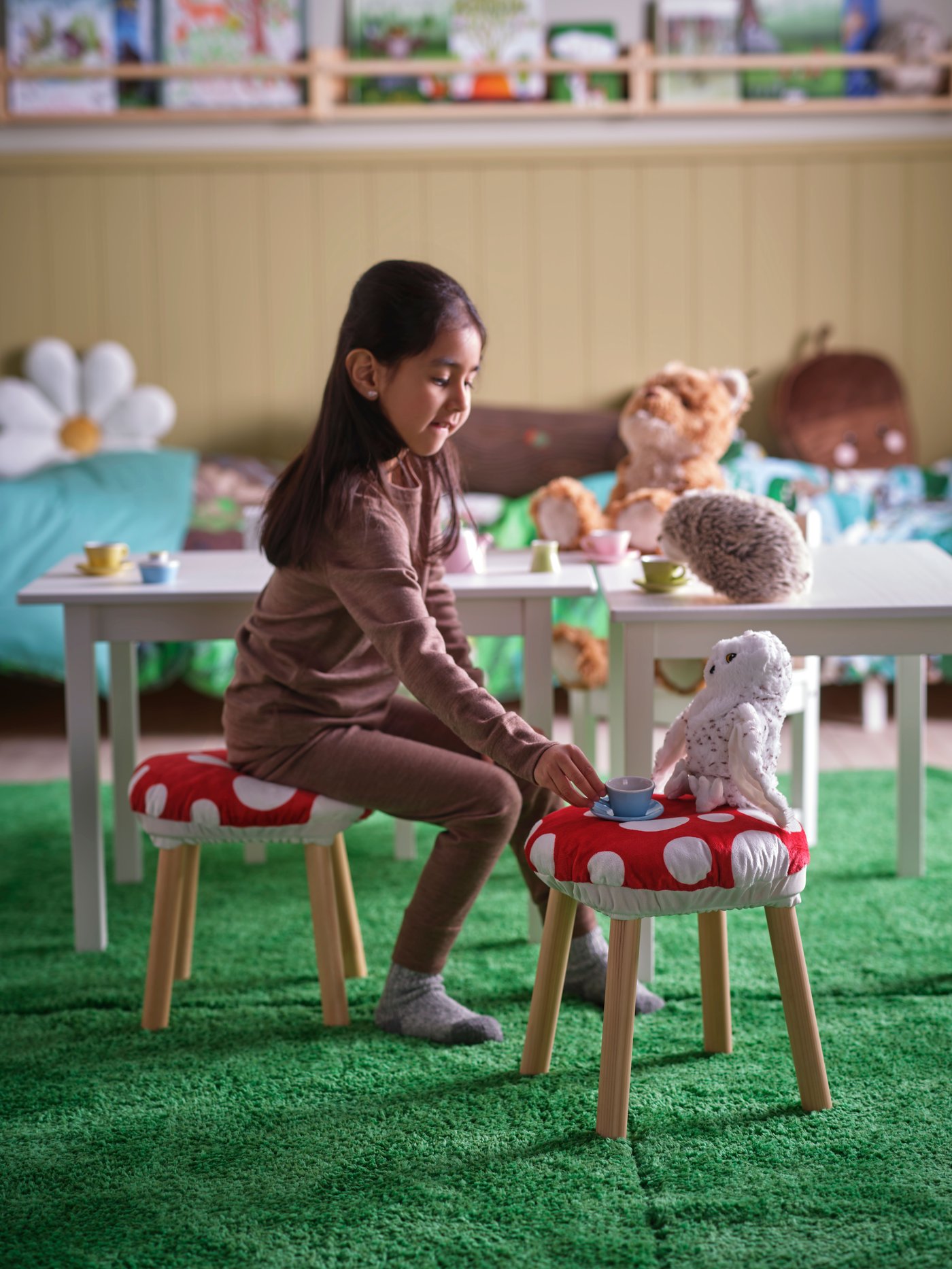 A child in a room surrounded by different products of the SKOGSDUVA collection. She is sitting on a small table surrounded by red and white toadstools. On one of the toadstools sits a soft toy owl that the child is offering a cup of tea.