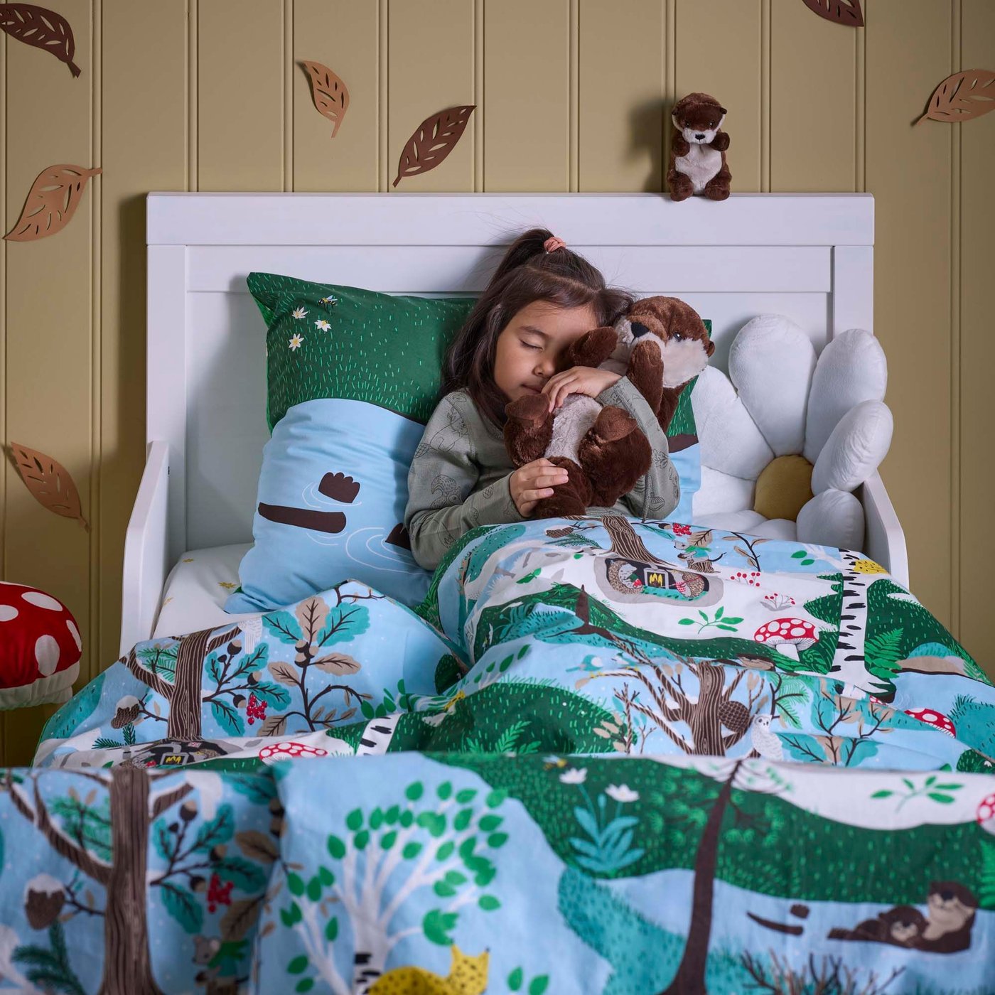A child hugging an otter with baby/brown beige SKOGSDUVA soft toy while lying in a bed dressed in SKOGSDUVA bedding.