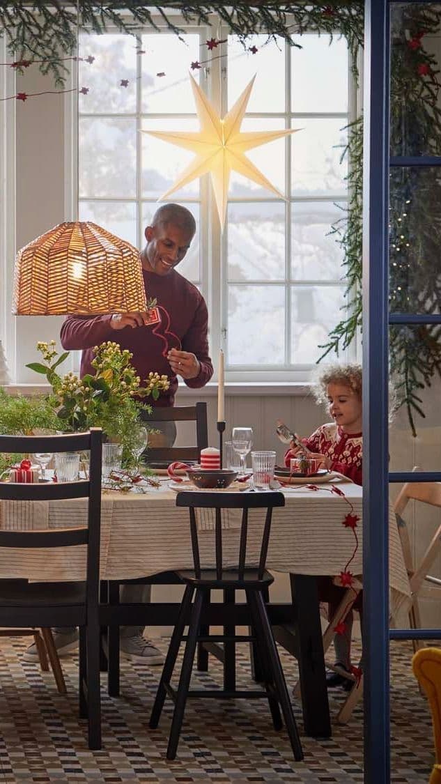 A dining area with a child and adult behind a dining table with a lit STRÅLA lampshade in the window behind them.