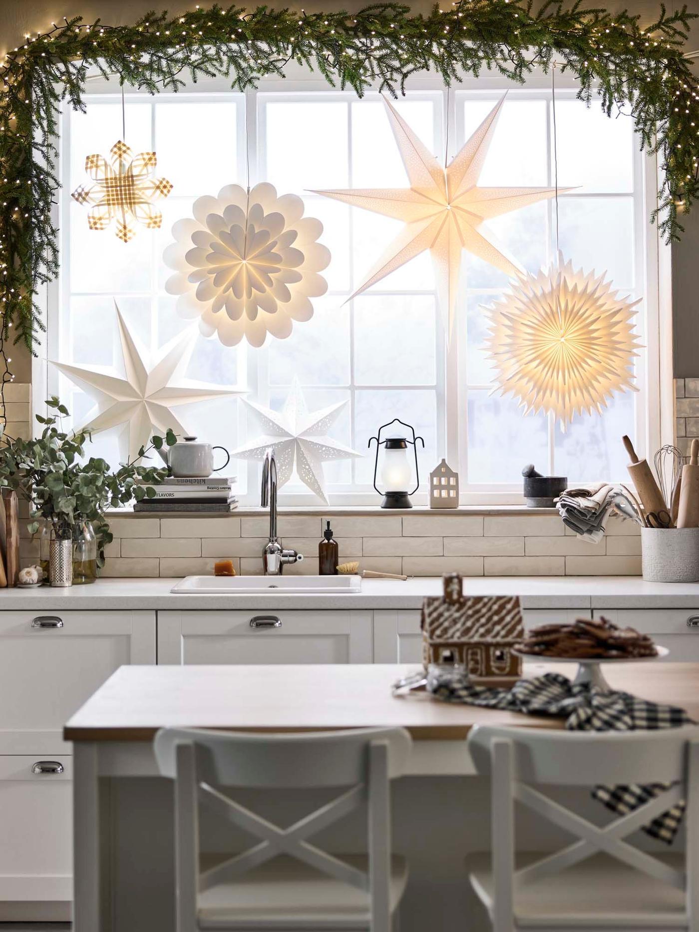 An illuminated assortment of STRÅLA lampshades in various star and flower shapes adorn the large window of a white kitchen and dining area.