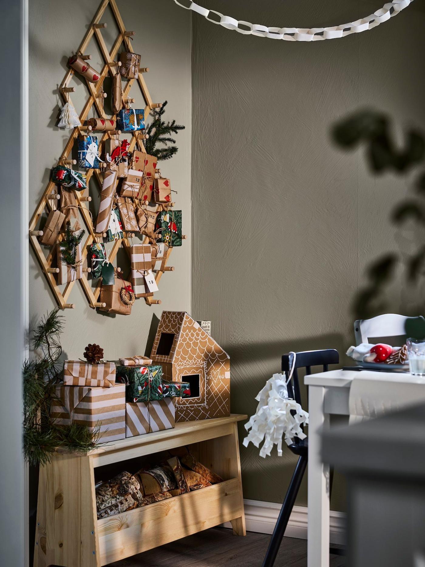 A VINTERFINT pine wall decoration filled with small gift bags and foliage hangs on a wall in a dining room, above a bench stacked with Christmas gifts.