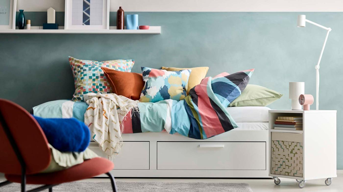 A bedroom roomset with a HEMNES daybed in focus, covered in colourful SVALSJÄRTAR bed linen, next to a chair and VIHALS bedside table.