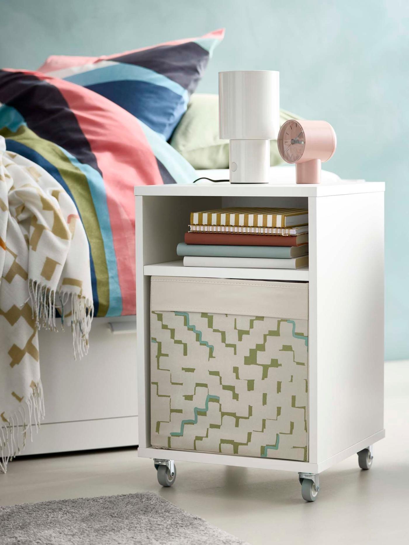 A VIHALS bedside storage table with a clock and lamp on top, next to a HEMNES daybed with colourful bed linen.