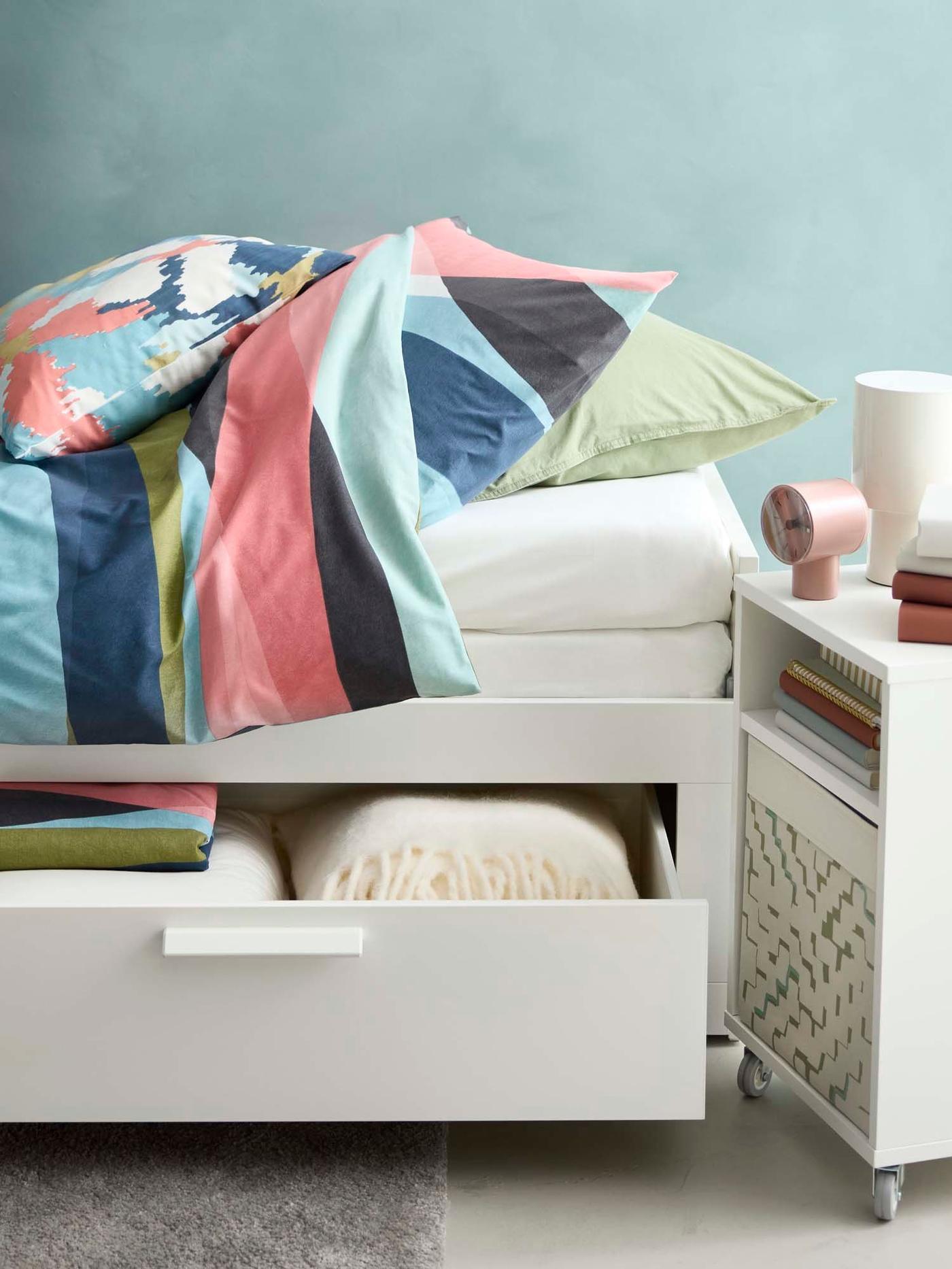 A HEMNES daybed with its underbed storage drawer open, filled with bed linen. The daybed has a colourful duvet on top.