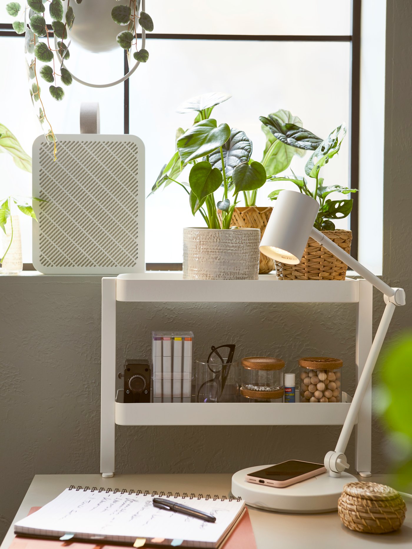 A desk beneath a window filled with plants. The desk is accessorised with a white lamp and shelves for a fresh, tidy feel.