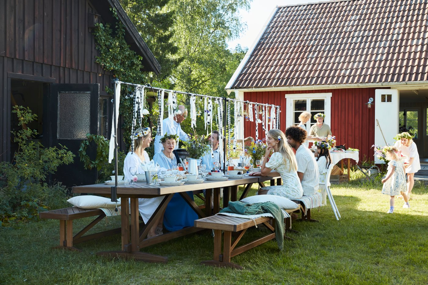 A group of people enjoy a midsummer party in a rural garden setting, with a traditional Swedish red house in the background.