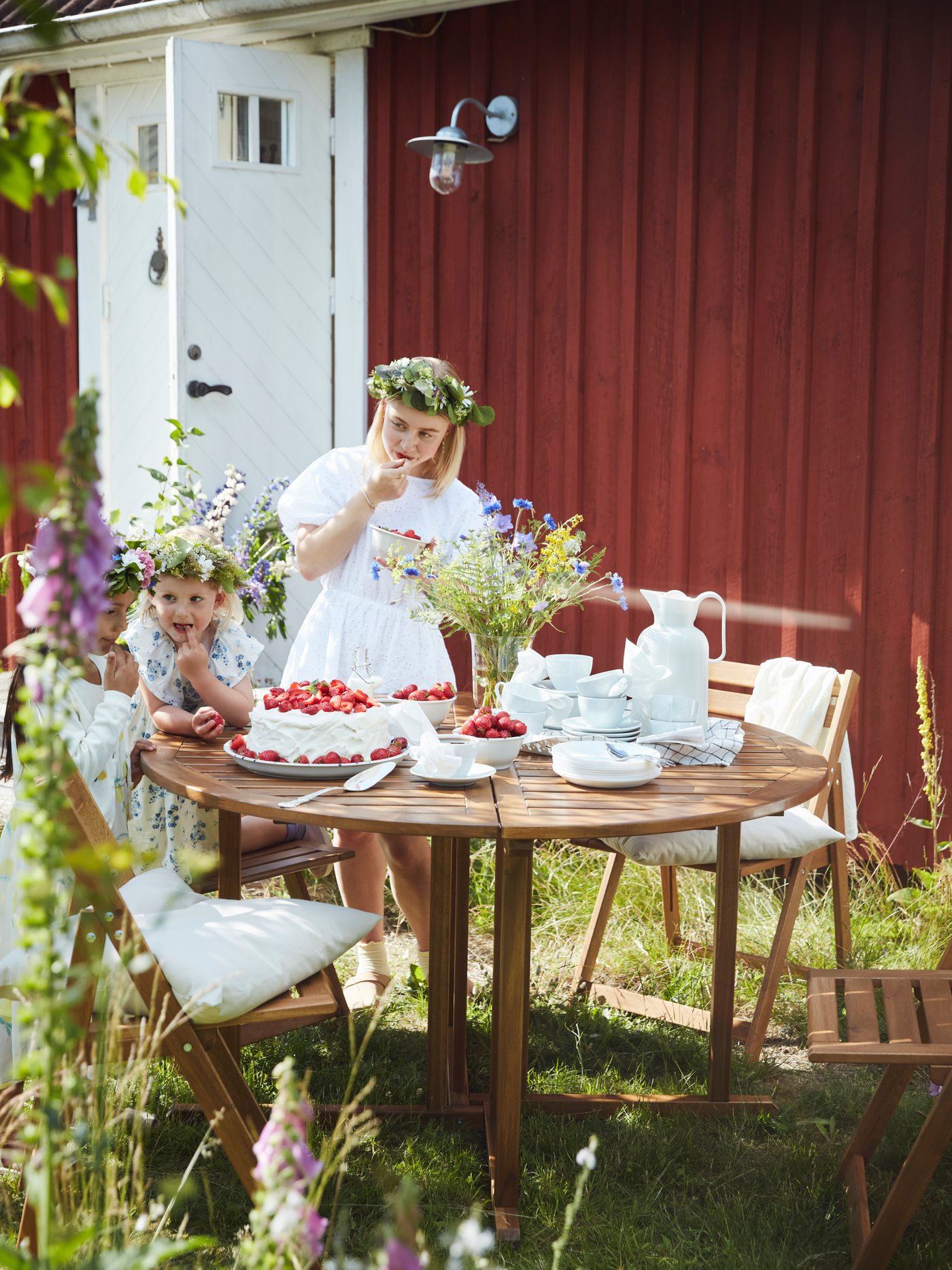 Three children sit and stand around a round NÄMMARÖ table outside on sunny day. They wear flowers in their hair and enjoy cake and fruit.