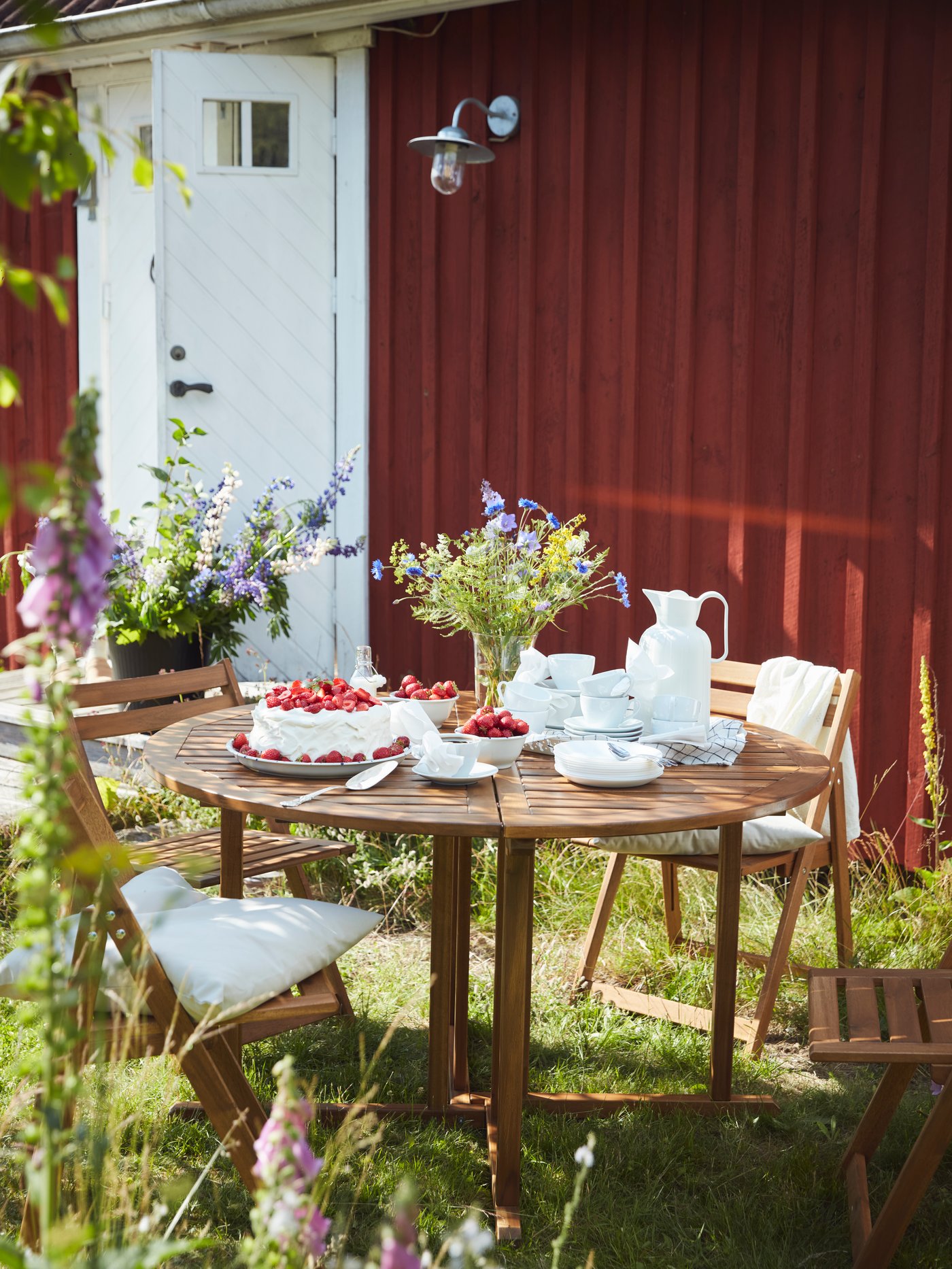 A round table and chairs in a rustic setting, prepared for afternoon tea, with cups and saucers and a strawberry cream cake.