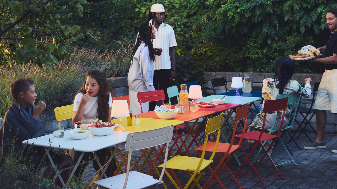 A group of people socialising outdoors around a row of SUNDSÖ tables in chairs in a rainbow of different colours. 