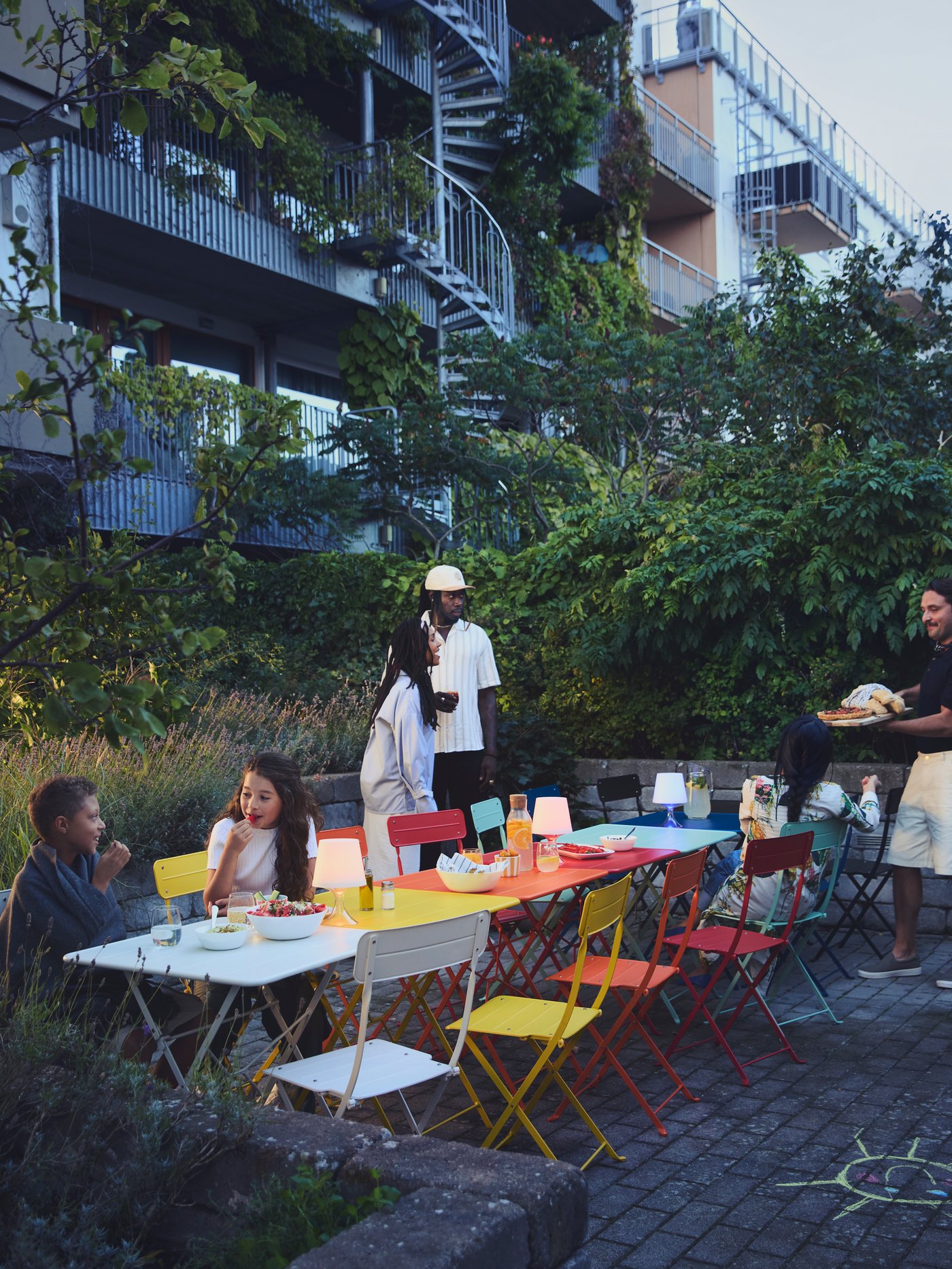 A group of people sit and stand, chatting around a long row of colourful tables and chairs, outside an apartment block. 