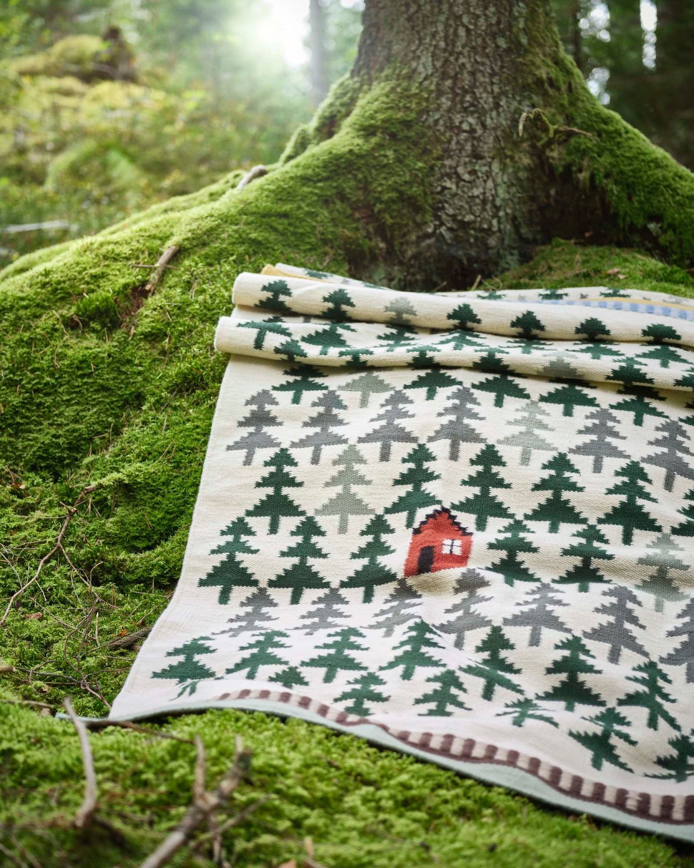 A DVÄRGGRÄS rug featuring a pine tree pattern with a little red house, is laid out on the mossy floor of a forest at the trunk of a tree.