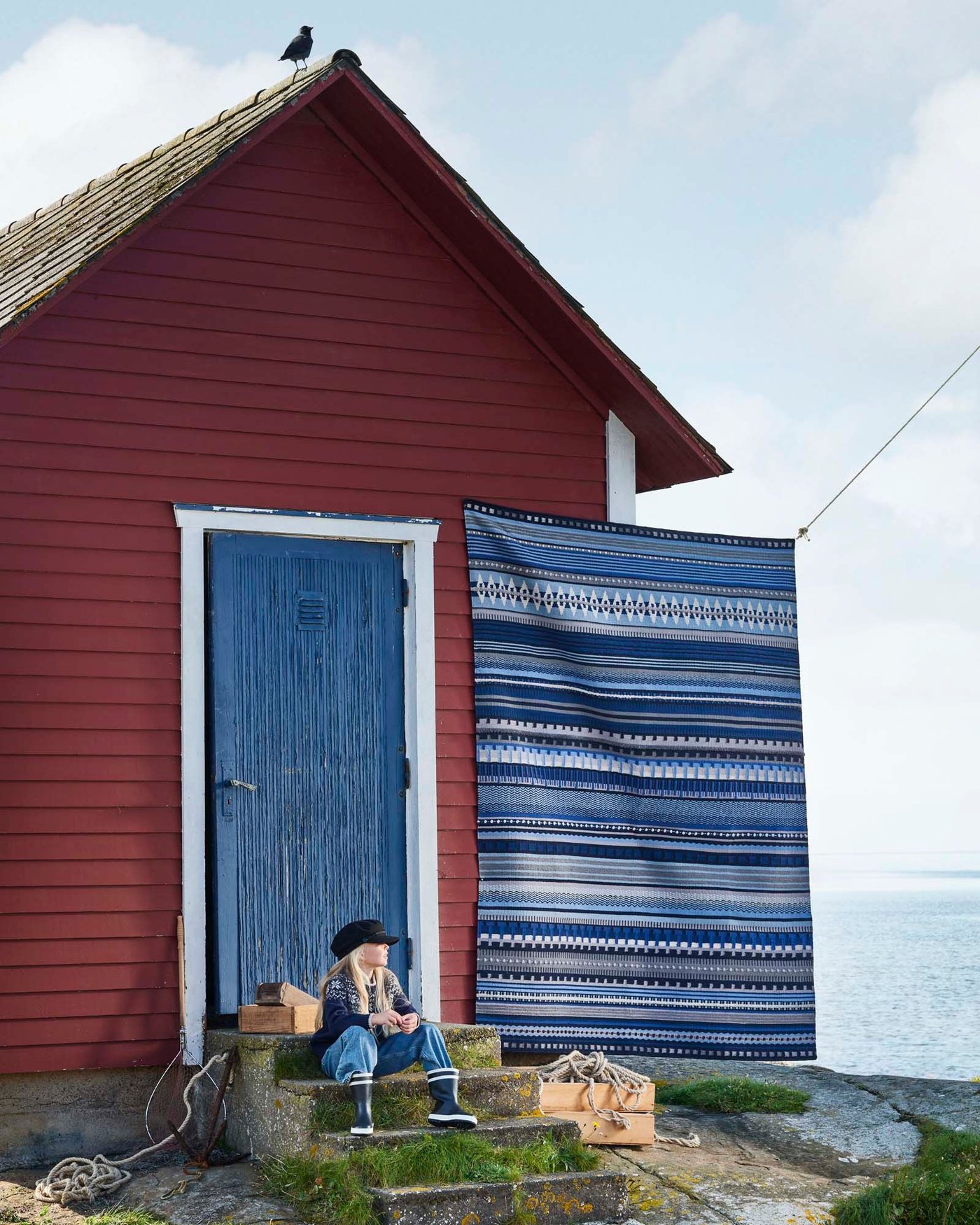 A child sits on the steps of a falu red fisherman's hut with a blue door. A HARSVANS rug hangs on a line next to the hut.
