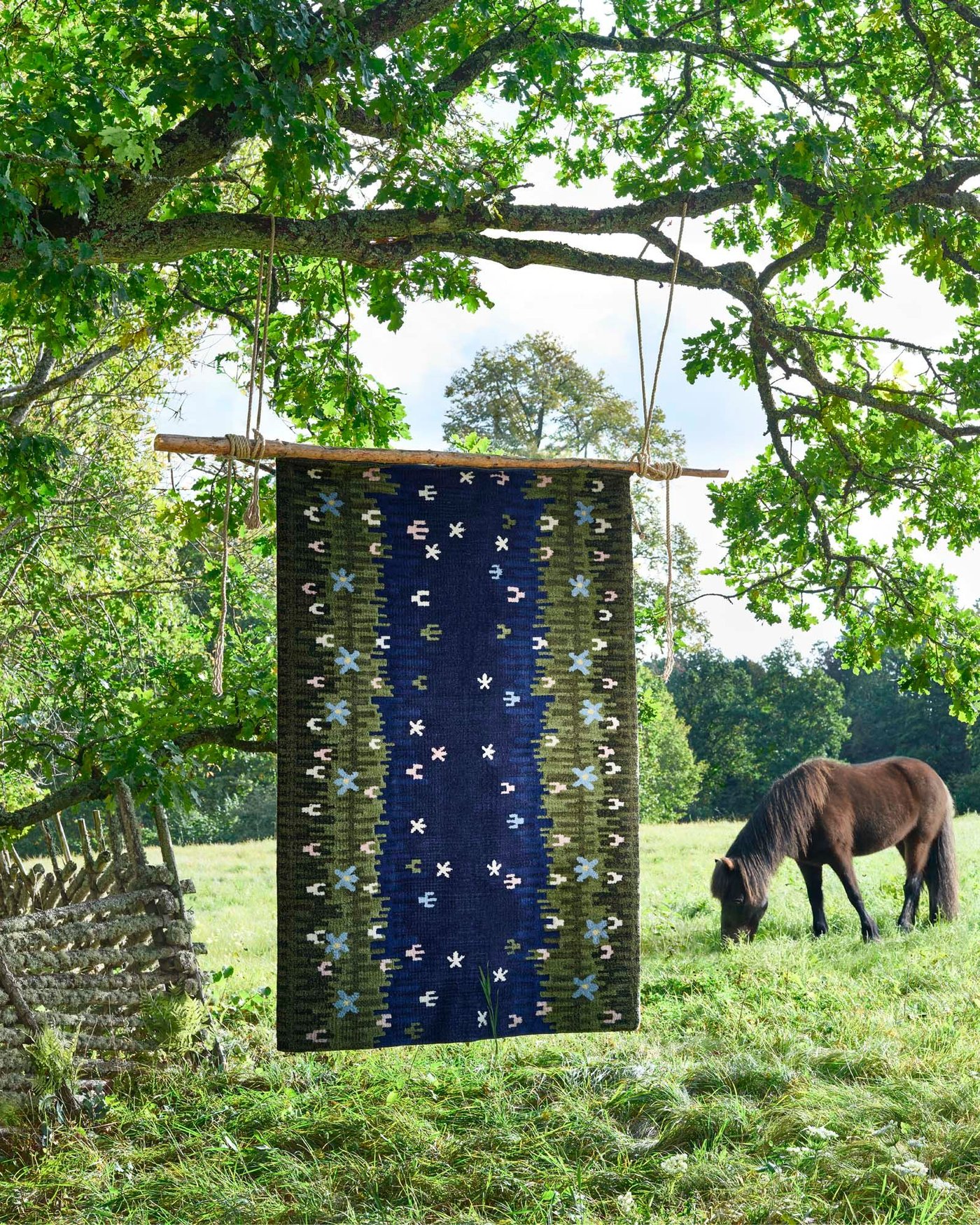 A GRODDSVINGEL rug hangs from a tree in a meadow. In the background, a horse is grazing.